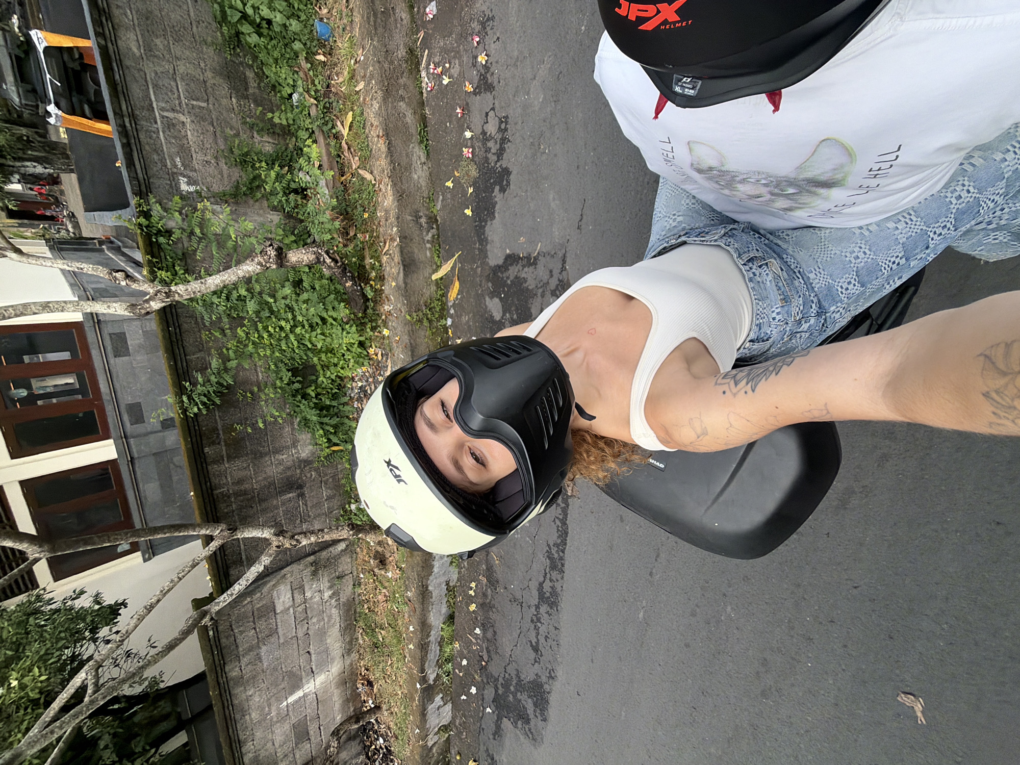 A first-person perspective shot captures a woman wearing a light cream motorcycle helmet with a black face mask, leaning back as if riding on the back of a motorcycle. She is positioned on a dark, possibly black, motorcycle seat. Her face, visible through the helmet's opening, is turned towards the camera, with her eyes looking sideways. She wears a white tank top and light blue denim jeans with a diamond pattern. Tattoos are visible on her right arm and shoulder. Beside her, a person wearing a white t-shirt with a graphic of a cow's head and text that reads "ONCE WE HELL" is seated, and a black motorcycle helmet with "JPX" branding is partially visible. The environment appears to be a street in North Kuta, Indonesia, with a textured gray stone wall lining the side of the road. Lush green vegetation grows along the wall and on the ground, and scattered white and yellow flowers are visible on the asphalt. Trees with bare branches arch over the scene, and a building with dark windows and a white facade is in the background. The road surface is asphalt, showing signs of wear and some wet patches.