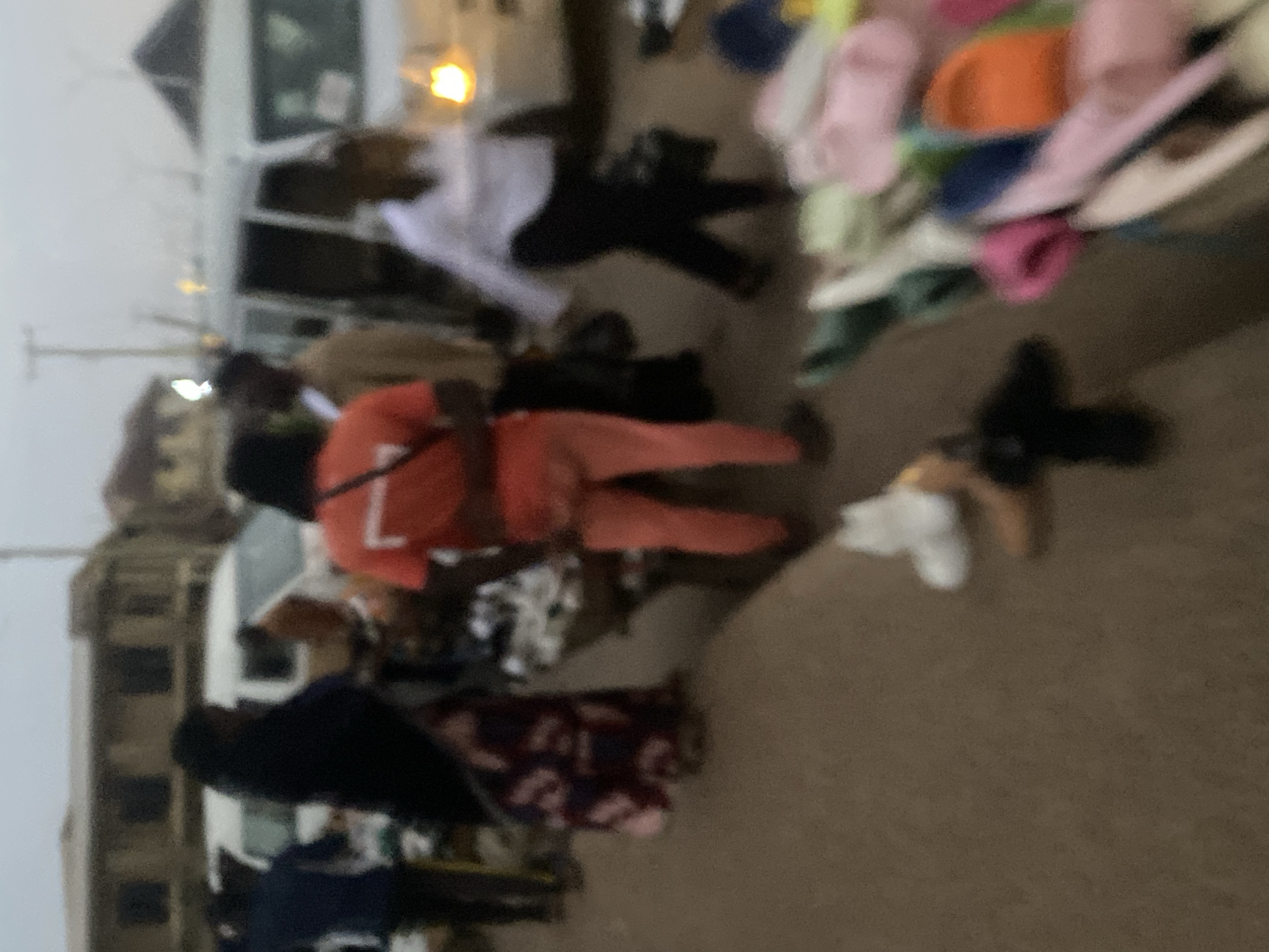 In Bukuru, Nigeria, a street scene unfolds with a shallow depth of field. A white minivan is visible, partially obscured by people. On the left, a person in a blue shirt and dark pants walks away from the camera. To their right, a person wearing a dark head covering and a patterned dress stands with their back to the viewer. In the center, a person in an orange t-shirt and orange pants is facing away from the camera. To their right, a person in a white shirt and dark pants walks away, carrying a dark bag. In the foreground, a collection of colorful footwear, including flip-flops and sandals in shades of pink, orange, and green, are displayed on the ground, some in a bin. The ground is paved with a rough, sandy texture, and the overall lighting suggests it is either early morning or late evening. Buildings with dark windows are visible in the background.