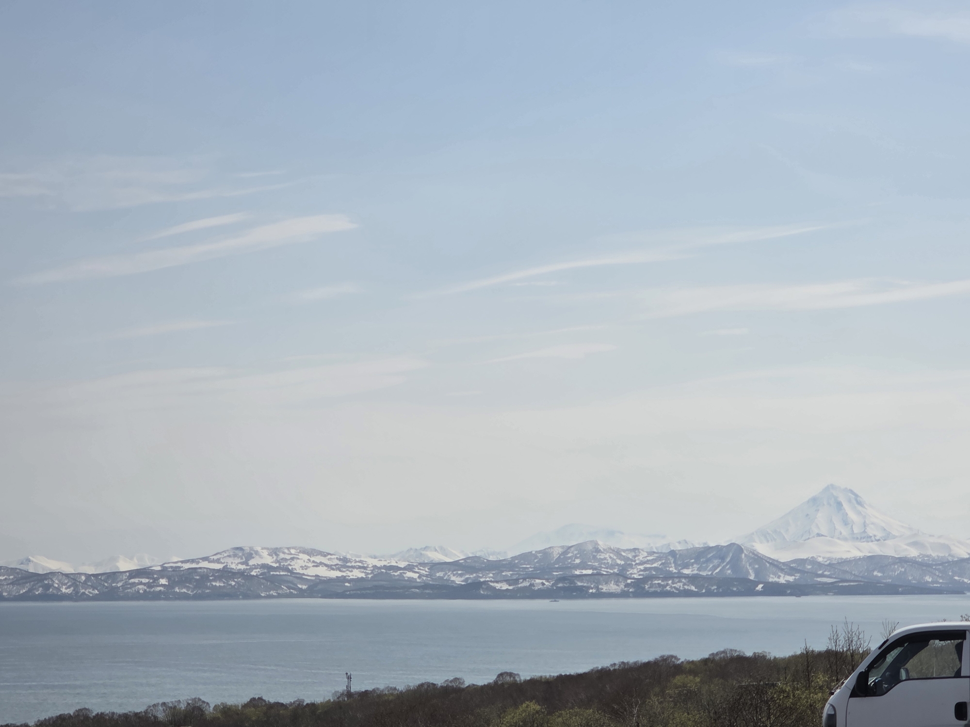 The image depicts a landscape with a large body of water in the foreground, beyond which are mountains covered with snow. The sky is light blue with thin, white clouds. In the lower right corner, a portion of a white vehicle is visible. The scene is observed from a high vantage point, suggesting a scenic overlook.