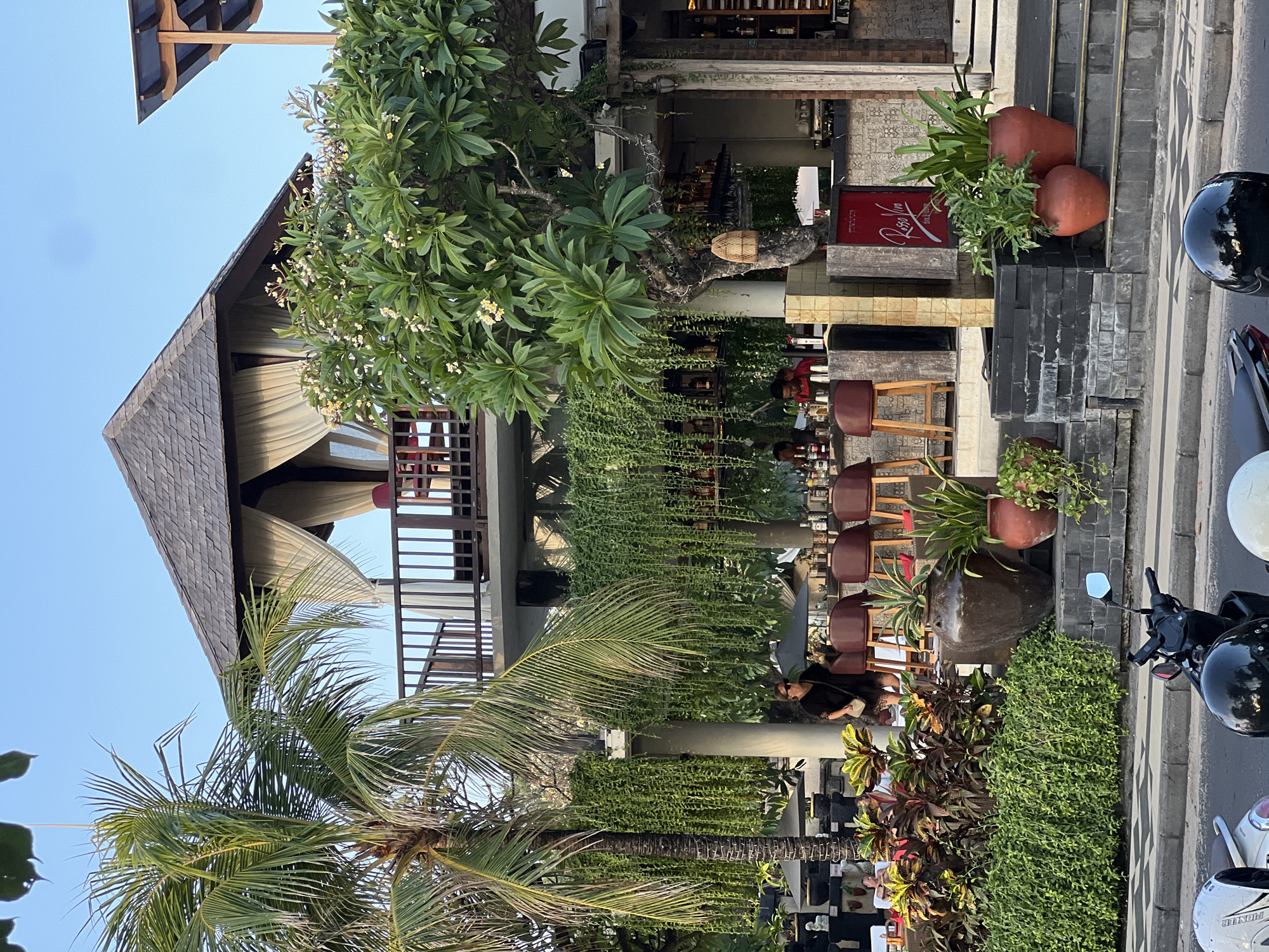 The image captures a street-level view of an outdoor bar and restaurant in Kuta, Indonesia, likely during late afternoon or early evening given the warm, natural lighting. The establishment features a multi-level structure with a thatched roof, open-air balconies, and lush greenery cascading down from the upper floors. A prominent palm tree stands to the left, with its fronds reaching towards the sky.
In the foreground, a paved sidewalk borders a street where several motorcycles are parked. The bar area itself is visible with high stools and patrons seated, some of whom appear to be staff members behind the counter. The bar is lined with shelves stocked with bottles. A woman in a black dress stands near the bar, looking towards the street. Large potted plants and decorative vases are placed on steps leading up to the establishment. A sign that reads "Rosso Viva" is prominently displayed. The overall atmosphere suggests a relaxed, tropical dining and drinking setting.