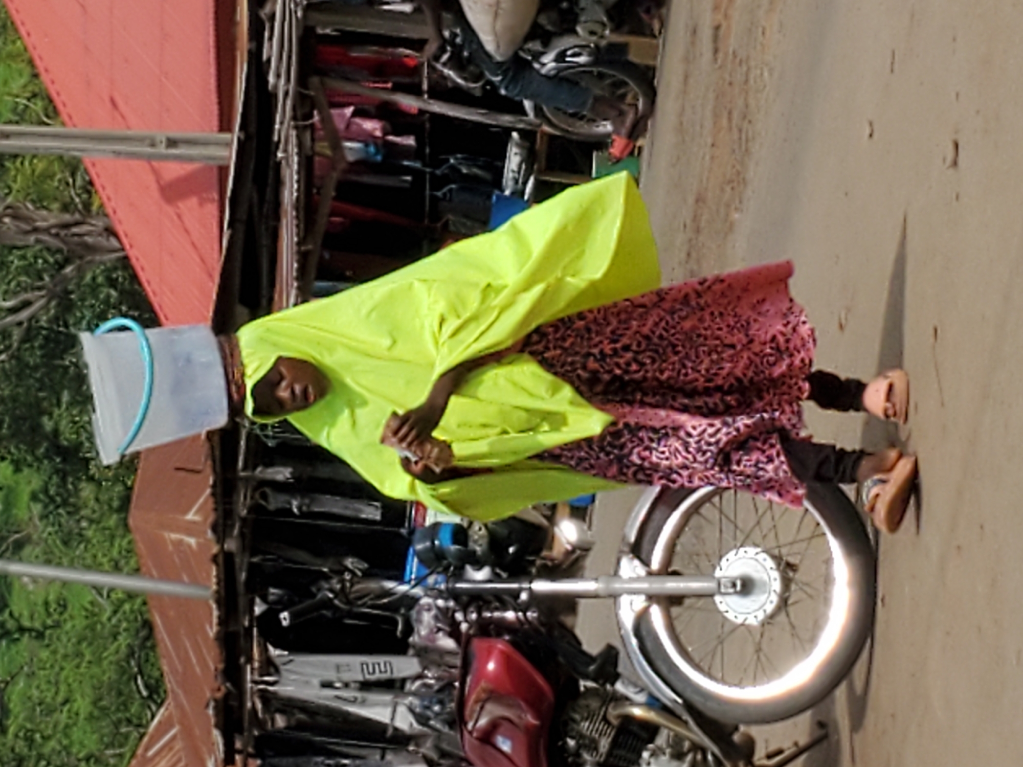 A woman wearing a bright yellow head covering and a pink patterned dress carries a bucket on her head while walking along a street in Mararaba, Nigeria. She is walking past a red motorcycle parked on the side of a concrete road. A building with a red roof and a collection of clothes hanging outside is behind her. A parked scooter can be seen in the background.