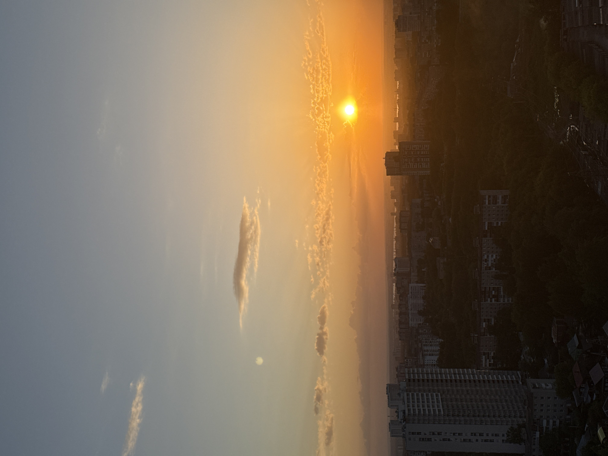 The image depicts a sunset over a cityscape. The sky transitions from light blue to orange as the sun approaches the horizon. Clouds are scattered throughout the sky, with some appearing as streaks or patches. The sun is a bright orb partially obscured by clouds. In the lower part of the image, buildings of varying heights dominate the urban landscape. Trees and other vegetation are visible amidst the buildings, creating a contrast between nature and urban development. The light from the setting sun casts a warm glow on the cityscape. The location is inferred to be Shirkovo, Russia, based on the prompt.