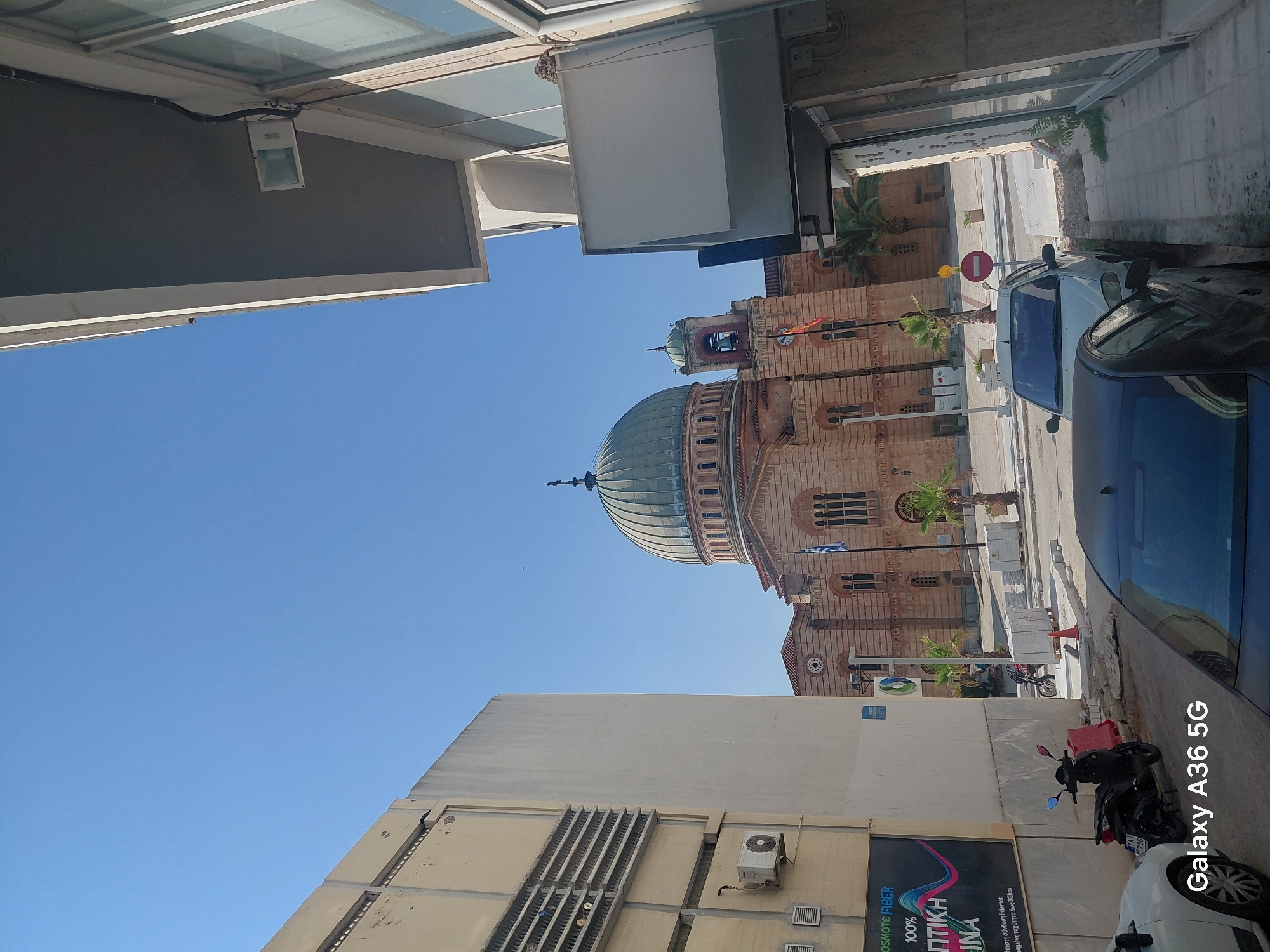 The image depicts an outdoor scene in the Municipal Unit of Aegio, Greece, featuring a large, ornate building with a prominent dome and smaller tower, likely a church or cathedral, constructed of reddish-brown brick with arched windows and decorative elements. In the foreground, to the left, is a multi-story building with light-colored walls and horizontal vents near the top, and an air conditioning unit attached to the lower portion. To the right, a modern building with large windows and a balcony overhang partially obscures the view of the sky. The street in front of the large ornate building is paved, with several palm trees planted along the sidewalk. A few cars are parked on the street, including a white sedan and a dark-colored vehicle in the immediate foreground. A black scooter is parked to the left of the white sedan. The sky is clear and blue. A sign with Greek text and an advertisement for "COSMOTE FIBER" is visible on the left-hand building. A Greek flag is flying near the ornate building. The overall impression is a sunny day in a city, with a mix of historical and modern architecture.