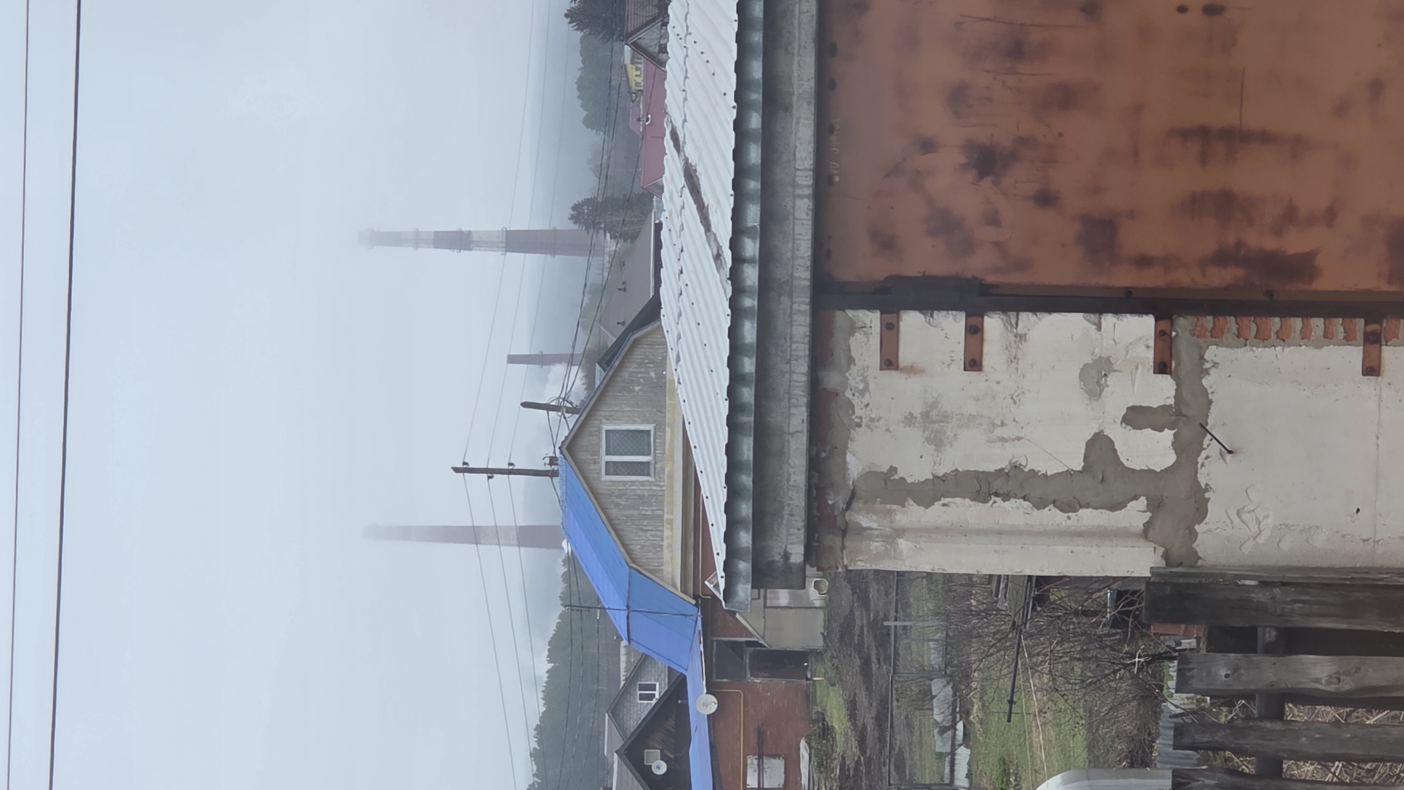The photograph shows a cityscape in Chaikovsky, Russia, shrouded in heavy fog. In the foreground is the corner of a building with a corrugated metal roof. Adjacent to this is a partially renovated wall with exposed brick and patches of bare concrete. Further back, several buildings with visible gabled roofs are discernible, some covered with blue tarps. In the distance, partially obscured by the fog, are tall industrial chimneys. Power lines run across the scene, adding to the industrial feel.