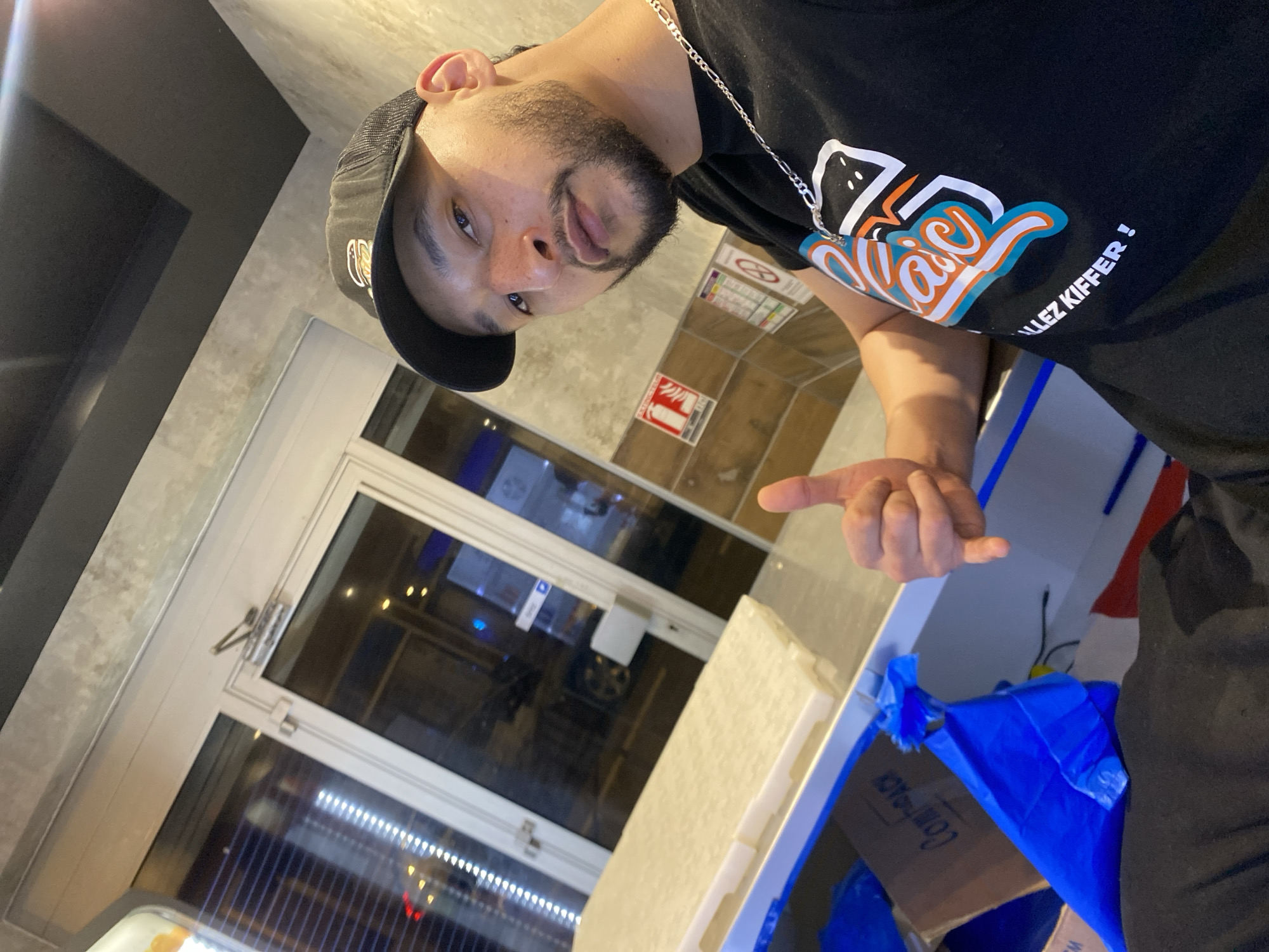 The image shows a man posing for a photo indoors, likely in the evening or at night, given the artificial lighting and reflections in the glass. He is seen from the waist up, wearing a black cap, a black t-shirt, and a silver chain. The man has a beard and mustache and looks towards the camera with a neutral to slightly smirking expression. His right hand is raised, with his index finger pointing towards the right side of the frame.
His black t-shirt features a colorful graphic design in white, orange, and blue, with stylized text that appears to be "Naïa" or similar, followed by the French phrase "ALLEZ KIFFER !".
The background includes a large glass door or window on the left, revealing reflections of lights and what appears to be a dark exterior. On the wall behind him, there are several signs. One sign displays a flame symbol and the word "EXTINCTEUR" (fire extinguisher), while another shows a red circle with a diagonal line, often indicating a prohibition. To the right, a white rectangular object, possibly packaging material, rests on a surface, next to a blue plastic bag partially covering a cardboard box with "Compack" visible on it.
The setting appears to be a commercial or public indoor space, aligning with the location context of Schiltigheim, France, given the French text on the shirt and signs.