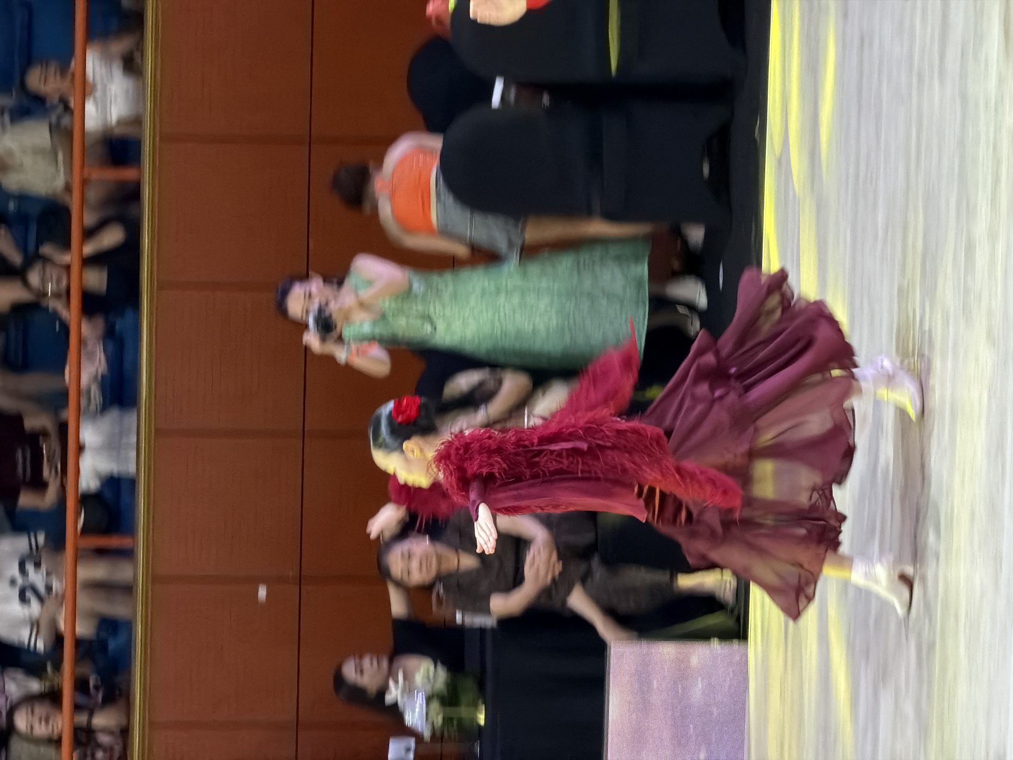 A woman in a red flamenco dress with feathered trim and a red flower in her hair is captured mid-twirl on a dance floor. Her dress billows around her as she wears white heeled boots. In the background, spectators are seated in a tiered audience, with some individuals standing and observing the performance. One woman in the middle ground is taking a photo with a camera. The environment appears to be an indoor venue with wooden paneling on the walls and a polished floor illuminated by yellow lighting.