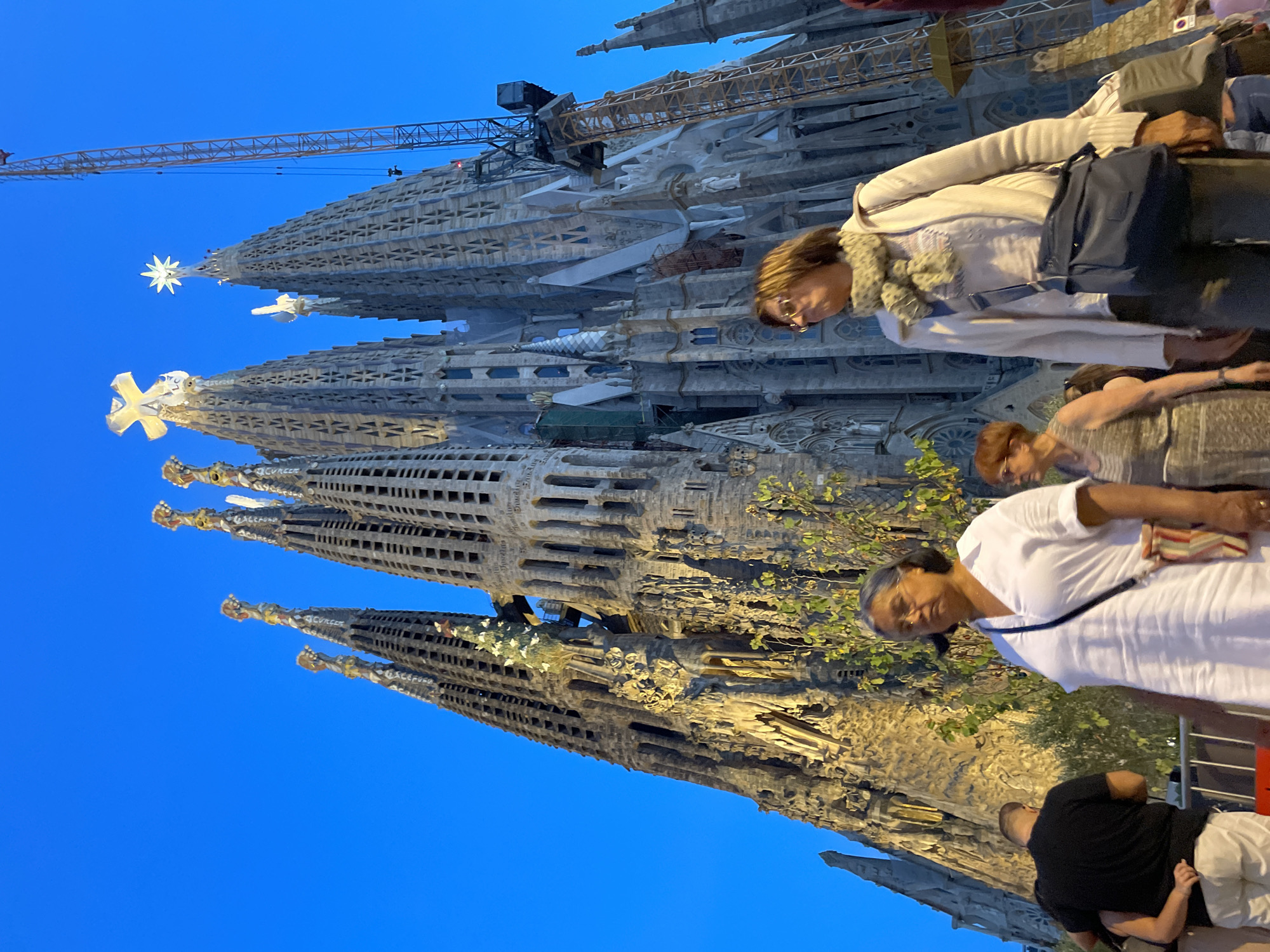 The image depicts the Sagrada Familia basilica in Barcelona, Spain, at dusk. The sky is a deep blue, and the iconic towers of the basilica are illuminated, showcasing their intricate architectural details. Several people are visible in the foreground, walking and standing. On the left, a person wearing a black t-shirt and white pants is seen from behind. To their right, a woman in a white dress with a striped bag slung across her body is walking towards the viewer. Further to the right, another woman in a beige cardigan and a patterned scarf, with a blue shoulder bag, is also walking. In the background, a construction crane is visible next to the basilica, indicating ongoing work. The overall scene is a blend of historical architecture and contemporary life, set against the backdrop of an evening sky.