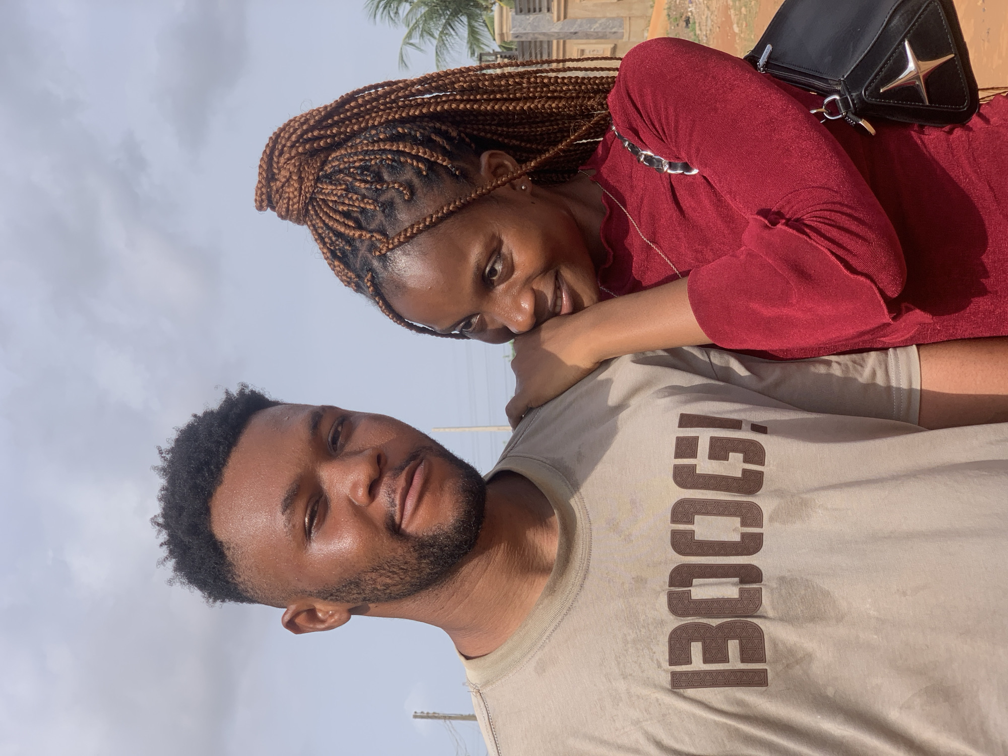 A photo captures a smiling dark-skinned woman with braided hair resting her head on the shoulder of a dark-skinned man with a beard. She is wearing a red, long-sleeved top and has a black purse with a silver detail on her shoulder. He wears a beige t-shirt with a dark print on the side. The photo was taken outside, likely in Ikorodu, Nigeria, given the presence of a palm tree and a structure with an unfinished appearance in the background. Overhead power lines are also visible. The subjects are in close proximity, suggesting a personal relationship.