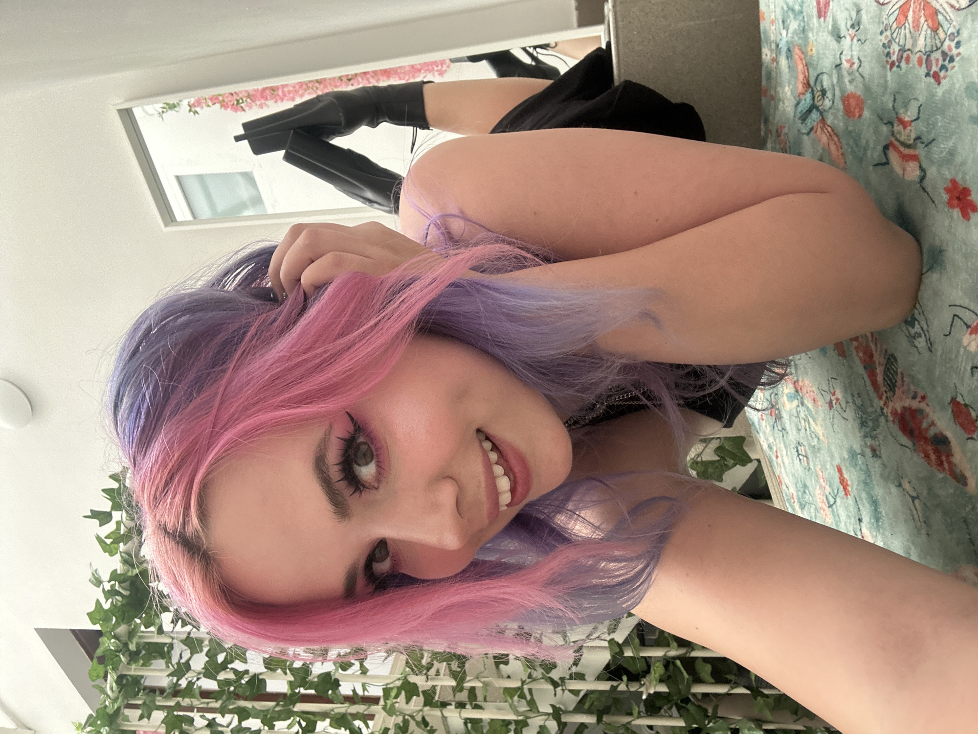 The image features a young woman posing indoors. She has striking, long hair dyed in vibrant shades of pink and purple, which cascades around her face and shoulders. Her eyes are accentuated with dramatic winged eyeliner and long, dark eyelashes, and she is looking directly at the camera with a wide, genuine smile, showing her teeth.
She is wearing a dark, possibly black, top or dress, and visible in the upper background are black knee-high or thigh-high boots, suggesting a stylish outfit. The woman appears to be lying down or leaning back on a surface covered with a light blue-green patterned textile, which features decorative motifs of butterflies, stylized insects, and floral elements in red, orange, and white.
In the background, above her head, there is a window or mirror reflecting an outdoor view, revealing pink flowers (possibly bougainvillea) and a white building, indicating natural daylight. Below her, green ivy vines are arranged decoratively, draped over a structure that might be a trellis or shelf.
The overall scene conveys a cheerful and playful mood, primarily through her smiling expression. The image is oriented dynamically, suggesting a selfie or a shot taken from above. The time of day appears to be daytime due to the natural light. No visible text is present in the image. The location context is the City of Zagreb, Croatia.