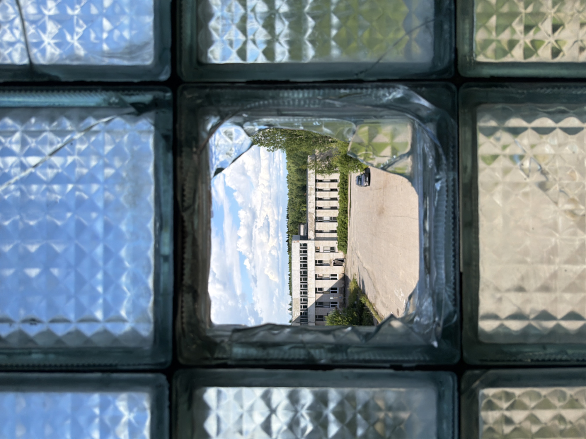 An exterior shot captures a broken glass block window in Nikolsky, Russia, offering a view of an abandoned building. The building, a long, light-colored structure with numerous windows, appears to be a former industrial or institutional facility. It is surrounded by overgrown vegetation and a paved, empty courtyard. A dark-colored car is parked in the courtyard in front of the building. The sky above is partly cloudy with white cumulus clouds against a clear blue backdrop. The foreground is dominated by the textured glass blocks of the window, with one central block shattered, creating an irregular opening. The overall scene suggests a state of disrepair and abandonment.