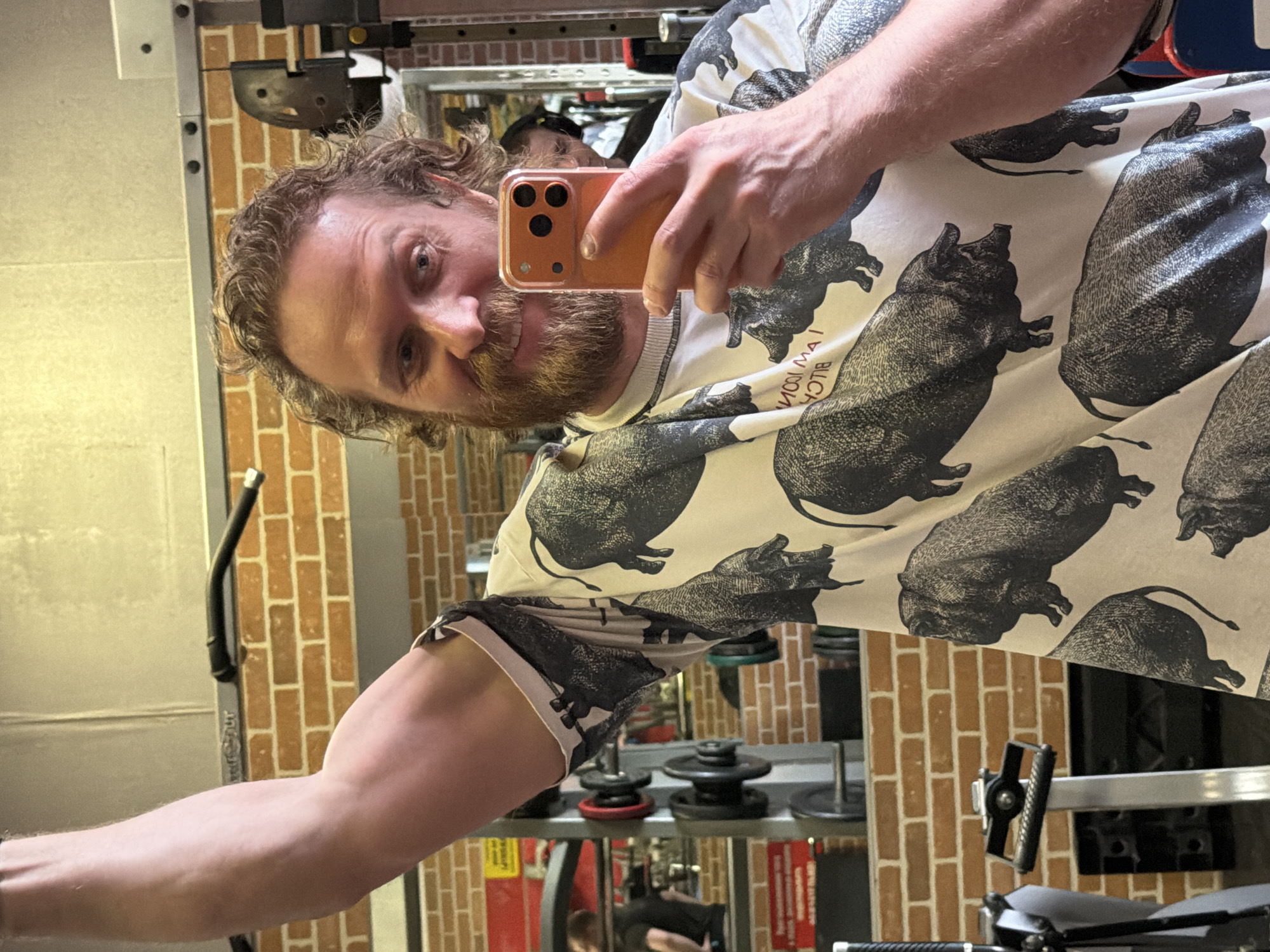 A man with curly brown hair and a ginger beard is captured in a selfie, looking directly at the camera with a slight smile and cheerful expression. He is wearing a light-colored t-shirt patterned with multiple black pig illustrations. Mirrored text visible on his shirt reads "I AM ICON / BLVCK." He holds an orange smartphone with a clear case and triple camera lenses in his right hand. His right arm is flexed, highlighting a bicep, suggesting a workout context. The background reveals gym equipment, including metal racks and weights. A distinct brick-patterned wall is also visible behind him. The image is rotated 90 degrees clockwise. The scene takes place indoors, likely during the day, in Saint Petersburg, Russia.