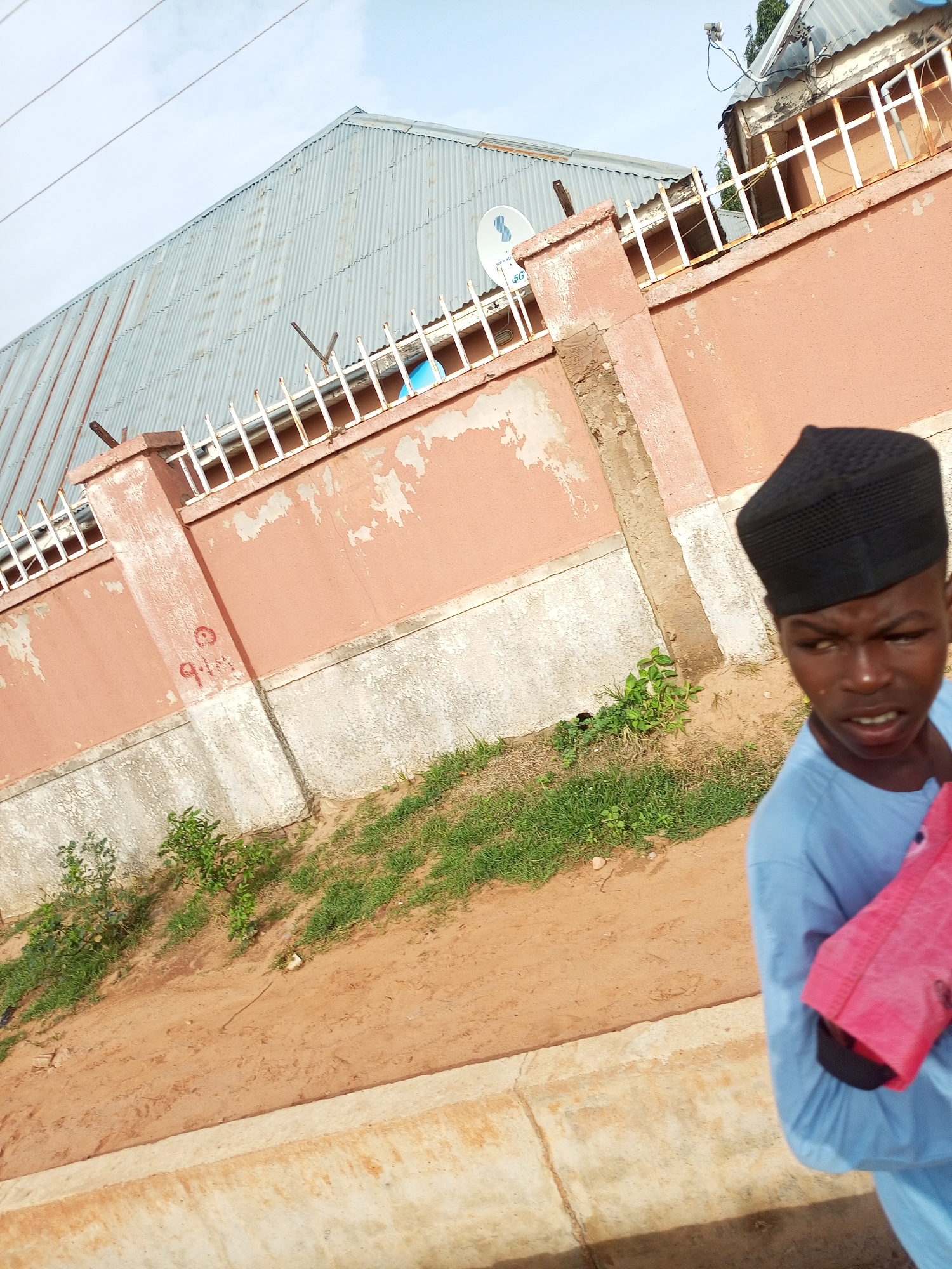 In Gombe, Nigeria, a young Black male wearing a light blue garment and a black kufi hat is shown from the chest up, looking slightly off-camera to his left with an open mouth and a surprised expression. He holds a bright pink cloth in his arms. He stands in front of a weathered, salmon-colored wall with white trim and a corrugated metal roof visible in the background. The wall is partly covered in moss and has sections of peeled paint. A white metal fence runs along the top of the wall, with some barbed wire. A satellite dish is mounted on the wall. The ground in front of the wall is sandy and covered with patches of dry grass and small green plants. The sky is overcast with some light clouds.