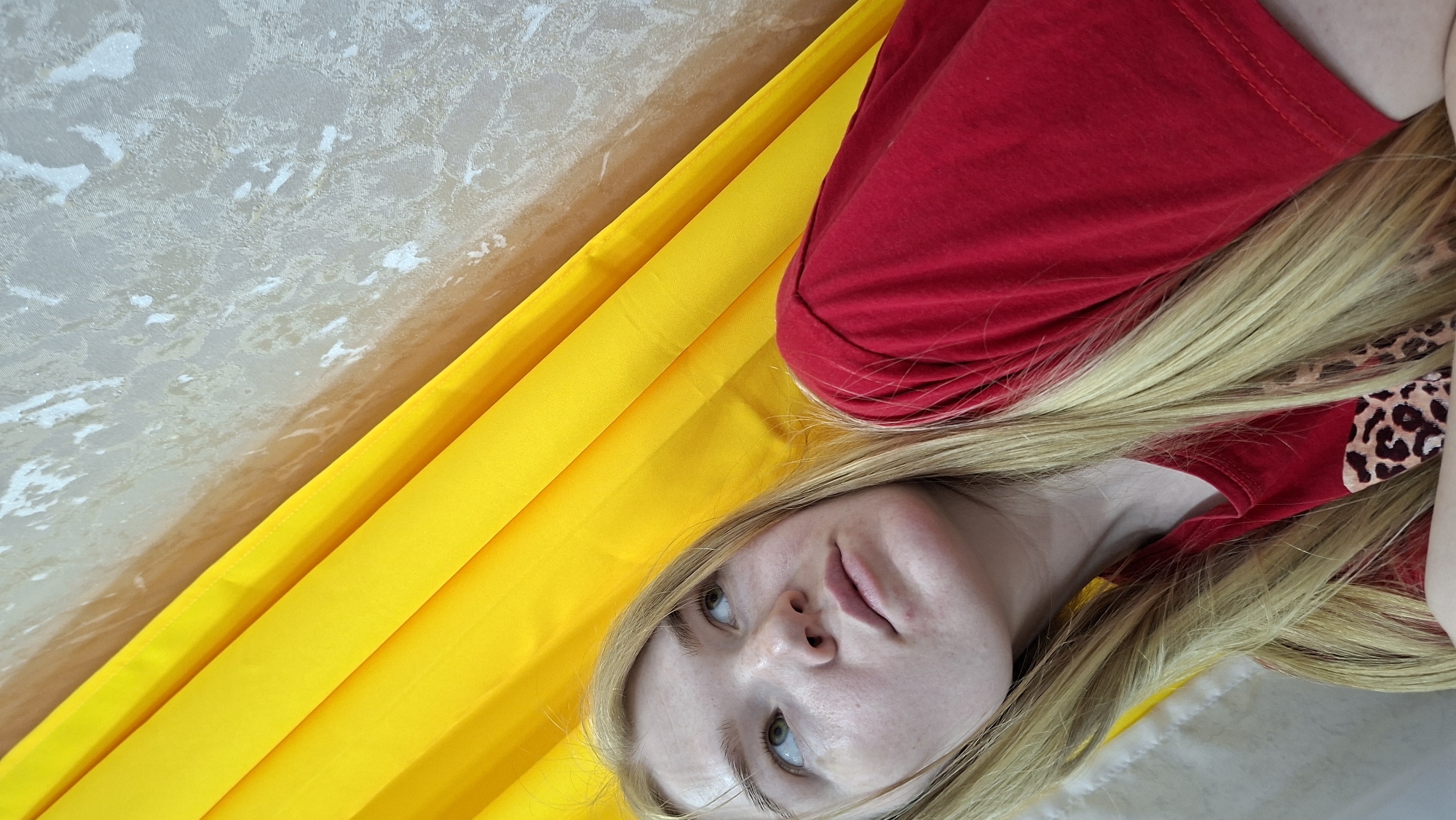 A blonde woman is lying on a surface partially covered with a yellow cloth. She is wearing a red top with a leopard print design. The background includes a textured, light-colored wall. No location information, including the city of Chaikovsky, Russia, can be confirmed from the image.