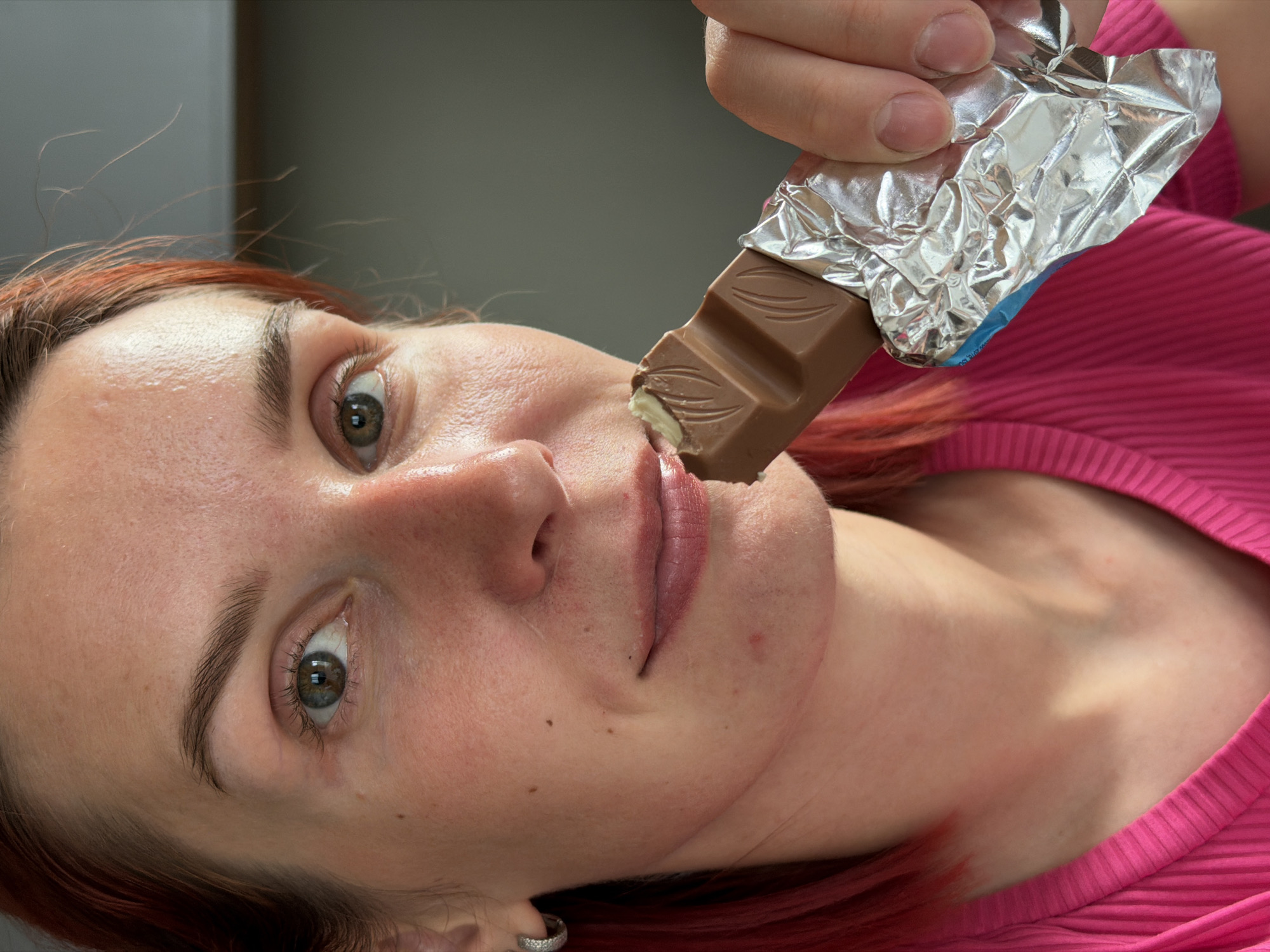 A person with red hair and fair skin is eating a chocolate bar. The chocolate bar is partially unwrapped and held in a hand. The person is wearing a pink shirt. The background is blurry and indistinct. There is no information in the image to indicate that this image was taken in Chaikovsky, Russia.