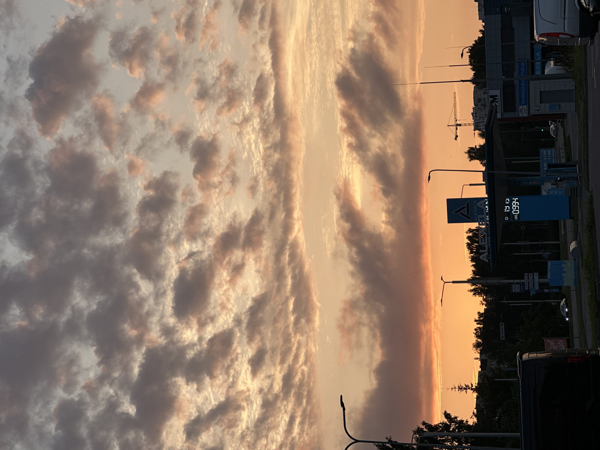 The image depicts a street scene in Tallinn, Estonia, during sunset. The sky is filled with a mix of gray and pink clouds, illuminated by the warm glow of the setting sun. In the foreground, a dark-colored van is parked on the left, and a white van is visible on the right. A gas station, branded "Alexela," is situated in the middle ground, with a digital price display showing "0.994" for LPG. A traffic light and street lamps are also visible. In the background, trees and buildings form a silhouette against the vibrant sky, and a construction crane is present. The overall scene suggests a typical urban environment transitioning into evening.