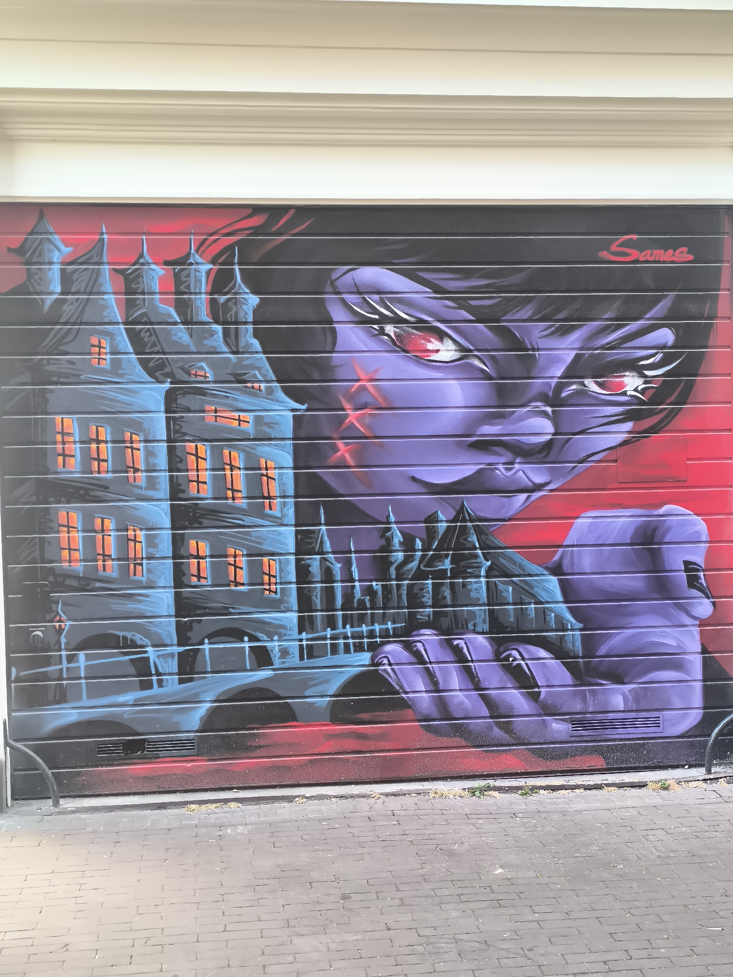 This image depicts a mural painted on a dark gray rolling security shutter. The mural features a stylized anime-esque character with purple skin and glowing red eyes, rendered in a dramatic and somewhat menacing style. The character's face is the dominant element, with sharp features and a piercing gaze. The background transitions from red on the right side to dark purple and black on the left.
To the left of the character, a detailed depiction of a gothic-style building, reminiscent of a castle or a haunted house, is painted. This building is rendered in shades of blue and gray, with illuminated windows showing a warm orange glow. Further to the left and slightly behind the main building, another, smaller structure of similar architectural style is visible. The character's left hand is positioned in the foreground, appearing to hold or interact with the smaller building, adding a sense of scale and narrative to the artwork.
The mural is signed with the name "Sames" in red lettering in the upper right corner. The security shutter itself is mounted above a paved area made of dark gray brickwork. A white, ornate cornice runs along the top of the shutter. The overall scene suggests an urban setting, likely on a street or alleyway where such security shutters are common. Without further context, a precise location within Amsterdam cannot be determined, but the artistic style and subject matter evoke themes of fantasy, horror, or gothic aesthetics.