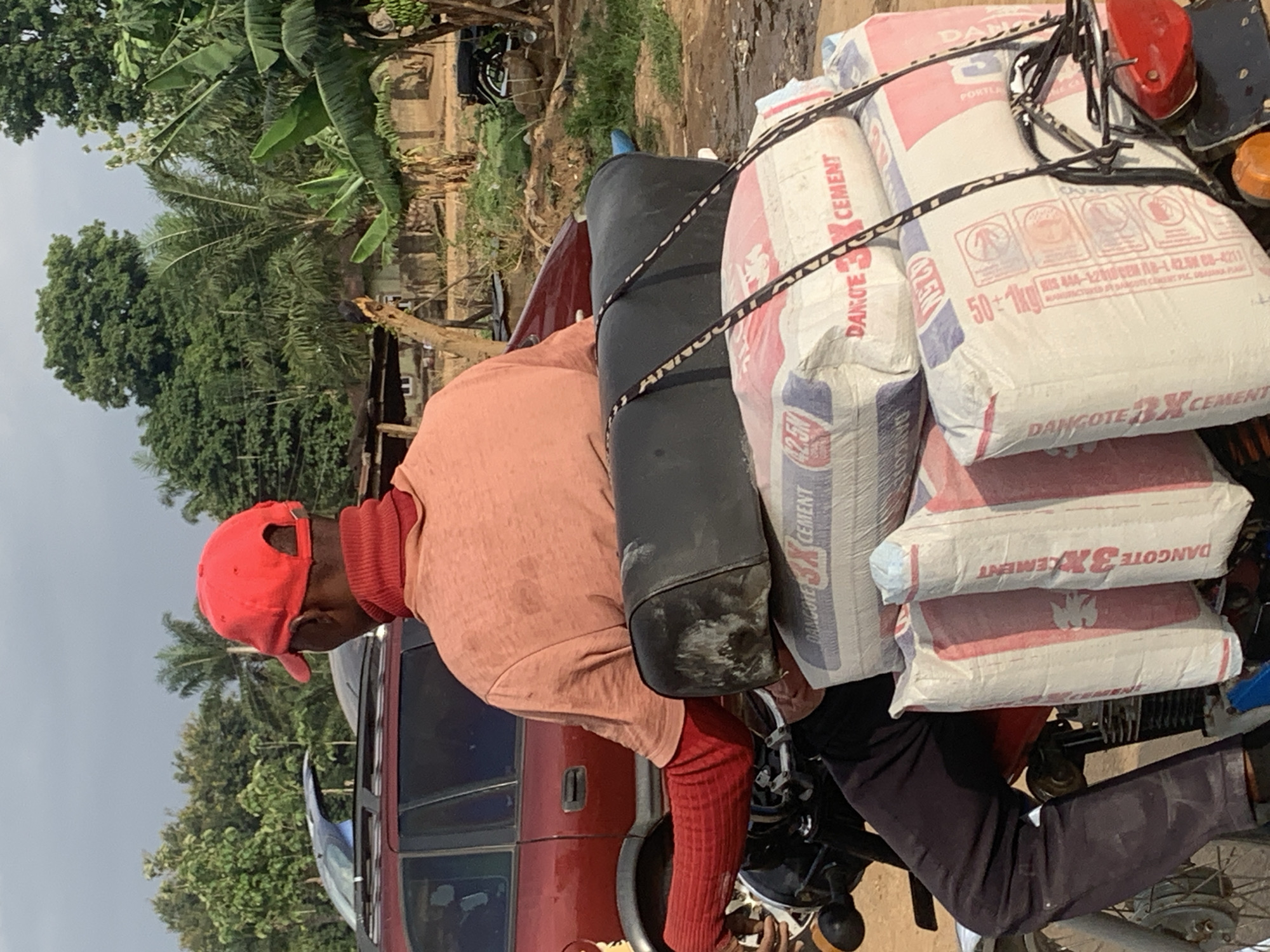 A man in a red cap and salmon-colored shirt is sitting on a motorcycle loaded with at least four bags of Dangote 3X Cement. The bags are stacked high and secured with black straps. The motorcycle is parked next to a red vehicle. The surrounding environment includes trees and a hint of a building or structure in the background. The image suggests the scene takes place in a typical urban area. Maigizo, Nigeria is likely to be incorrect.