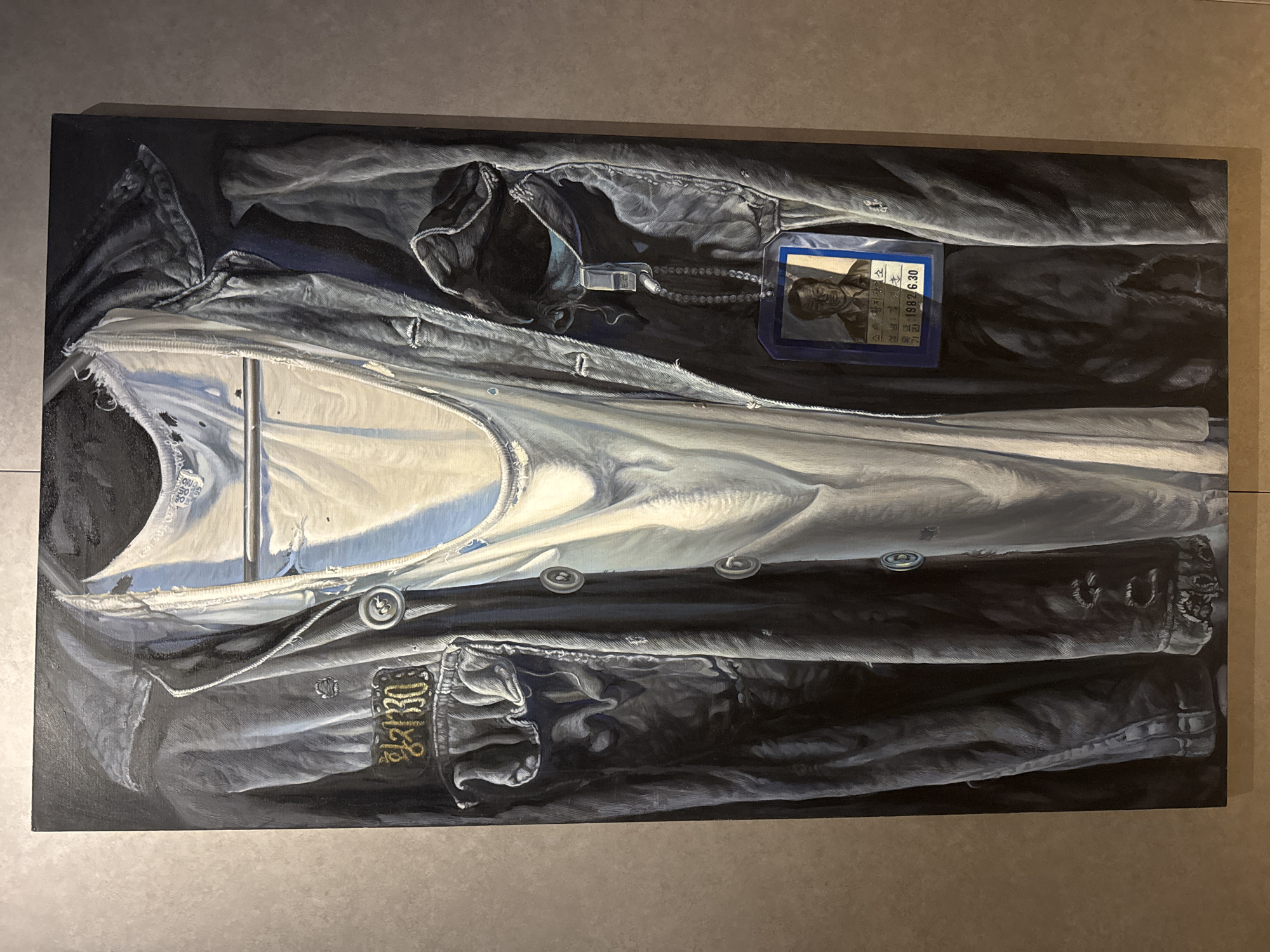 The image is a painting of a black leather jacket with a white shirt underneath. The jacket has buttons, pockets, and embroidered details on the sleeves. There is a blue tag with a portrait and Korean text attached to the inside of the jacket. The tag includes the date "1992.6.30". The painting is displayed against a neutral, light gray background, suggesting it might be hanging in a gallery or a home. There is no specific indication of the location being in Seoul, South Korea.