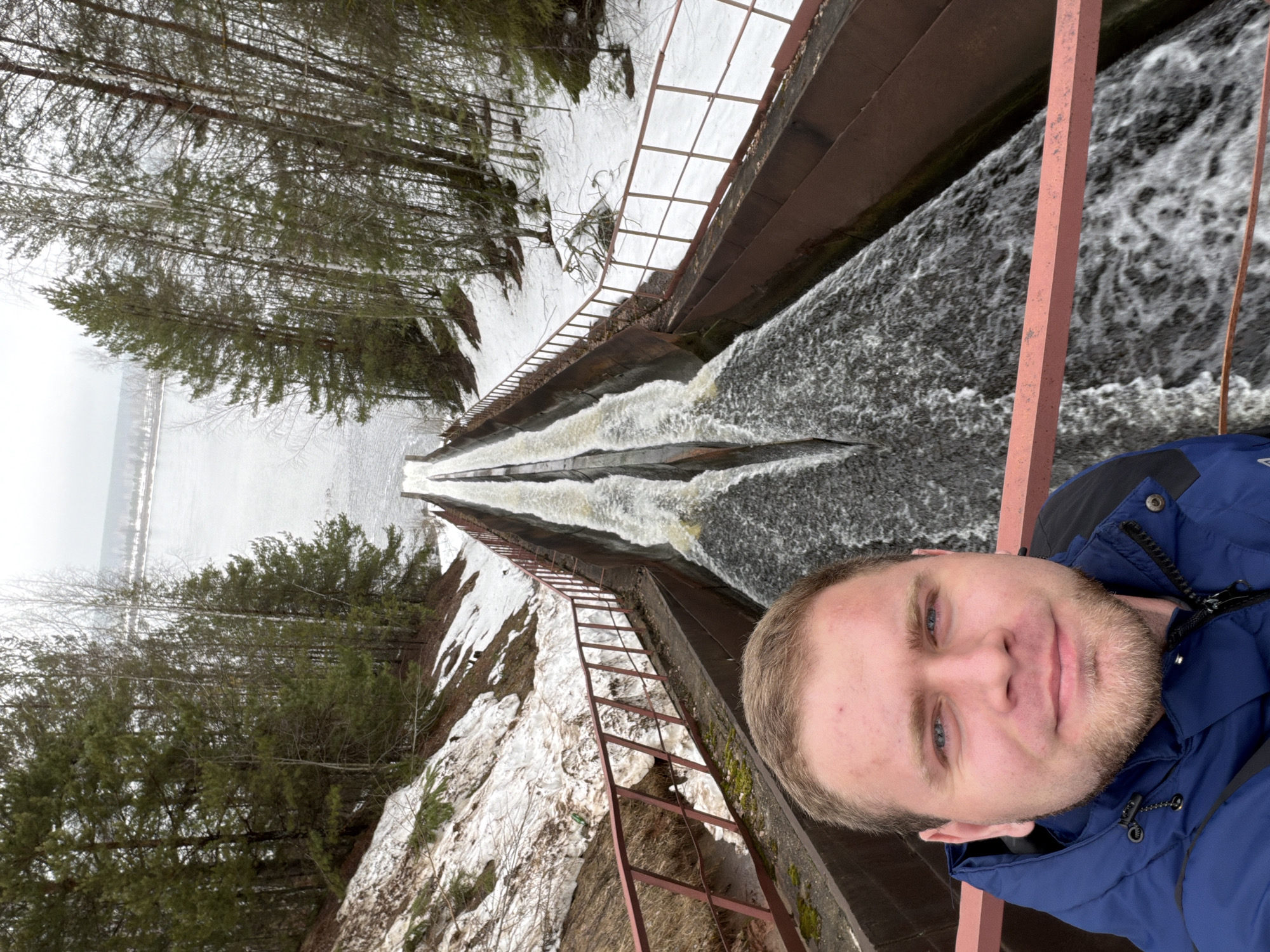 A man in a blue jacket is taking a selfie near a concrete aqueduct with water flowing through it. On the left side of the aqueduct, there's a steep embankment covered with snow and trees. The scene appears to be located in a rural or natural setting, potentially near Chaikovsky, Russia. A rusted ladder extends down the embankment next to the water. The sky is overcast.