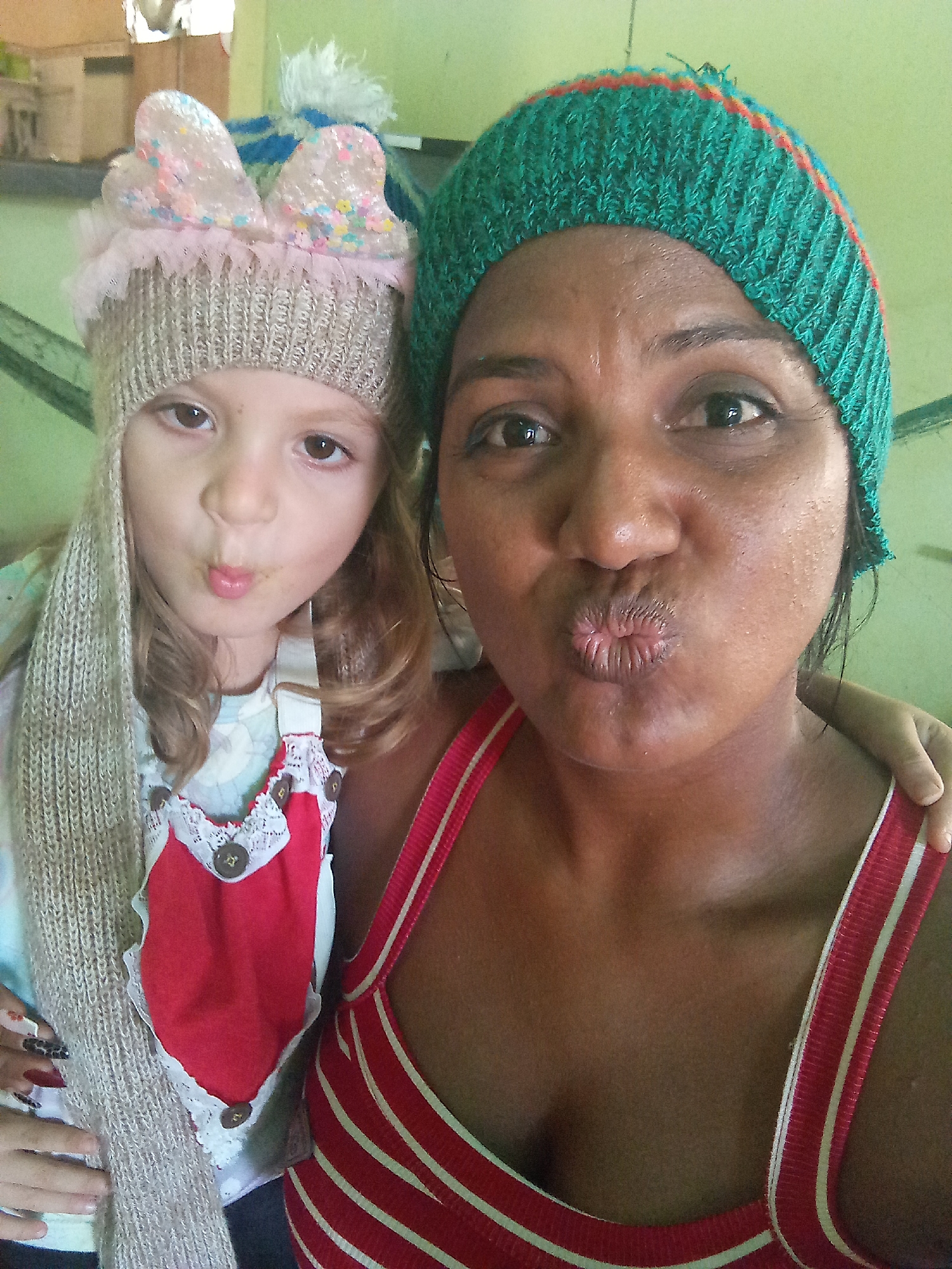 The image depicts two individuals, a child and an adult, posing closely together. Both are making a kissing face towards the camera. The child is wearing a beige knitted hat with decorative ears and a long scarf. The child also has on a red apron style garment with white trim and buttons. The adult is wearing a green and orange knitted hat and a red and white striped tank top. The background appears to be a room with light green walls and some furniture visible. It is not possible to confirm if the image was taken in the city of Altagracia, Venezuela, or elsewhere.