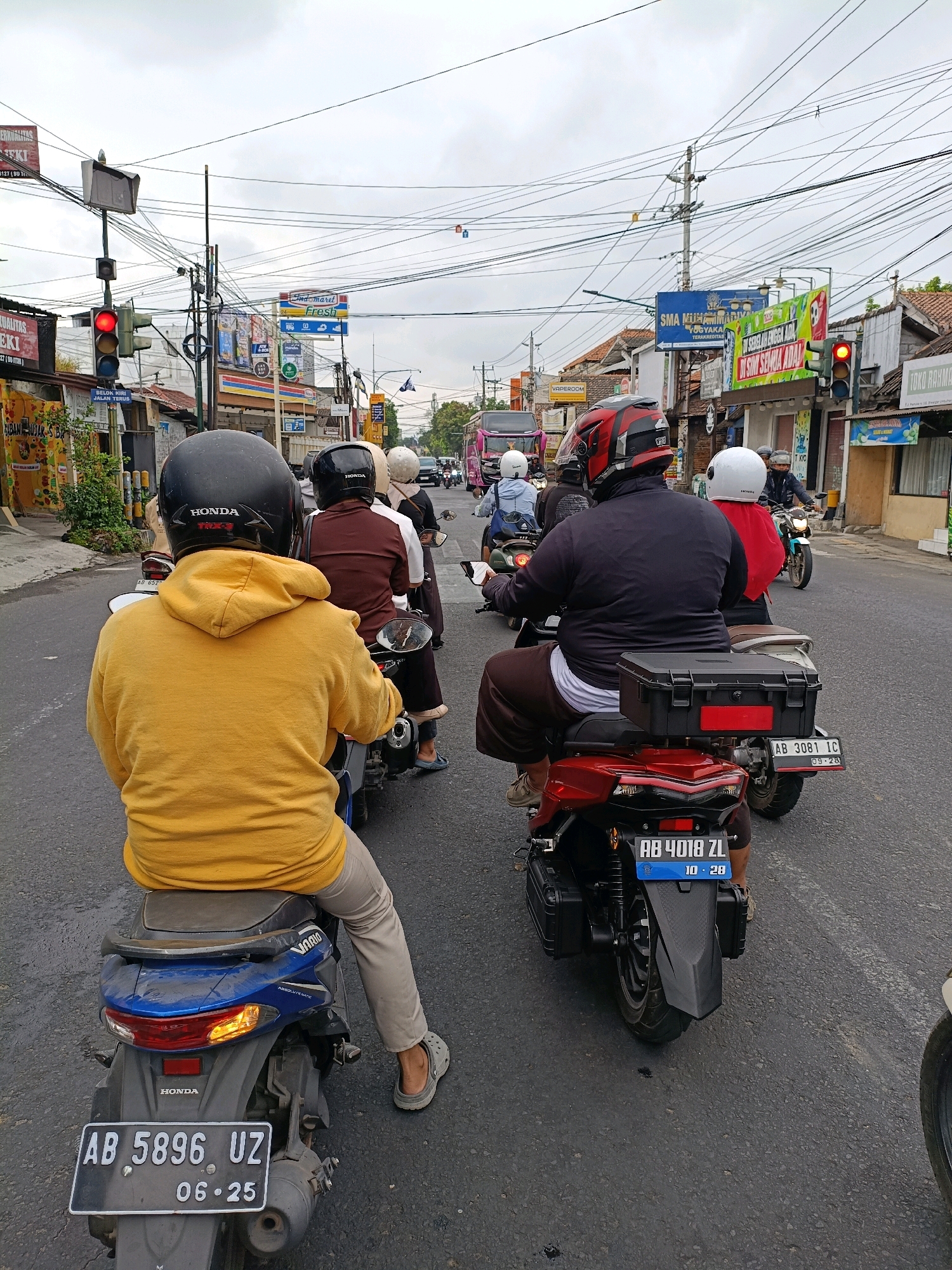 This image captures a busy street scene in Yogyakarta, Indonesia, likely during daylight hours under an overcast sky. A group of motorcyclists are navigating a two-lane road lined with various shops and signage, indicating a commercial area. In the foreground, a person wearing a yellow hooded jacket and a black helmet rides a blue motorcycle, their license plate reading "AB 5896 UZ 06.25". Several other motorcyclists are visible ahead, some with passengers, all wearing helmets. One motorcycle in the middle of the frame has a red rear and a black storage box on the back, with the license plate "AB 4018 ZL 10.28". The road is asphalt and appears to be in fair condition. Overhead, a complex network of power lines crisscrosses the sky, a common sight in Indonesian cities. Signs for businesses like "Indomaret Fresh" and what appears to be a school "SMA MUHAMMADIYAH" are visible. Traffic lights are also present, with one showing a red light. The overall impression is one of everyday urban life in Yogyakarta, with moderate traffic flow on a somewhat cloudy day.