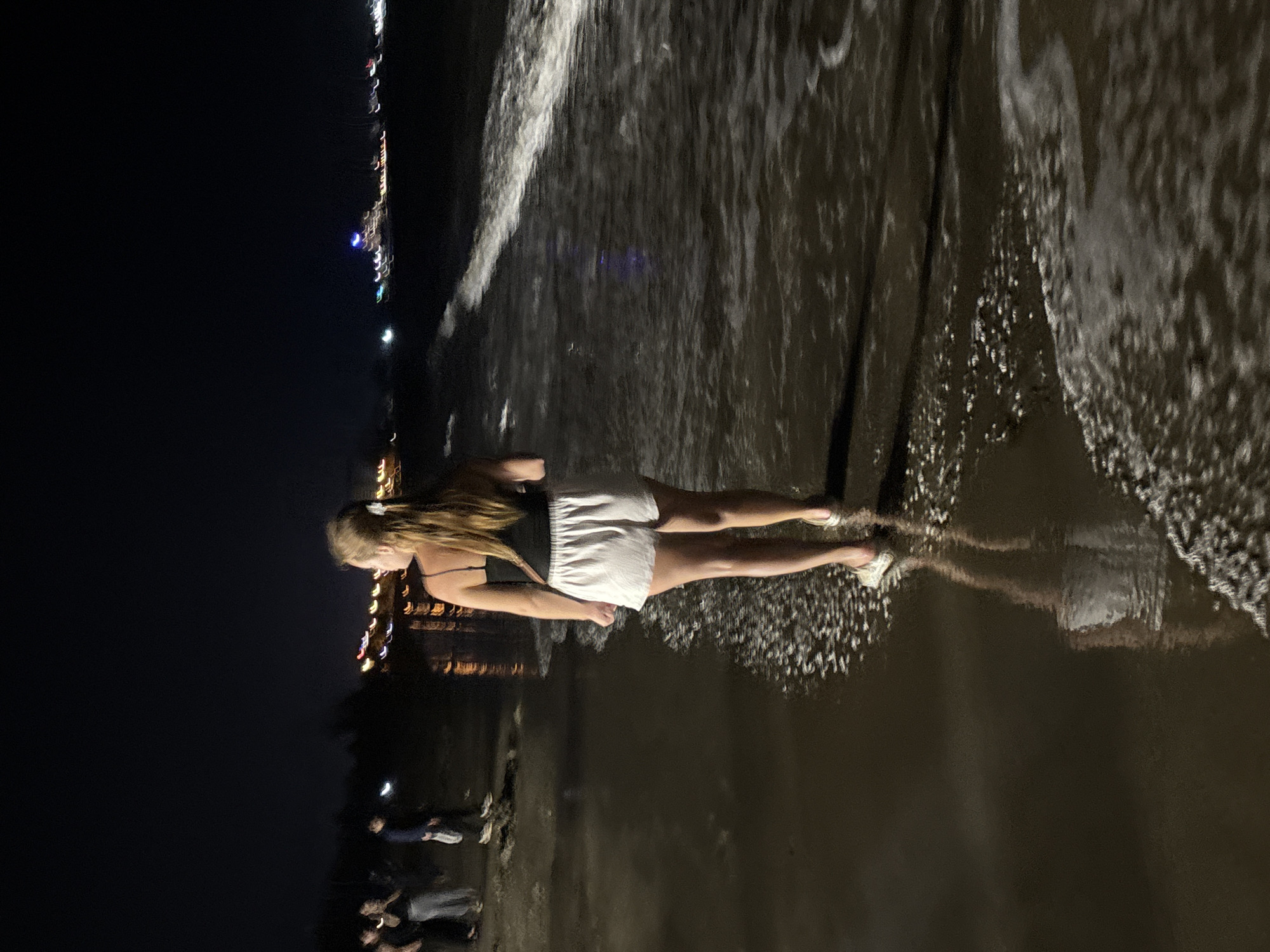 This vertical image depicts a nighttime scene at a beach or shoreline, set in Dong Hai Ward, Vietnam. The main subject is a young woman with long, light-colored hair, standing barefoot in shallow water or wet sand. She is wearing a dark top and white shorts, and is positioned slightly to the left of the center, facing partially away from the viewer and looking downwards.
White, foamy waves are visibly breaking around her feet, creating intricate patterns on the dark, reflective surface of the water or wet sand. A clear reflection of her lower body and legs is visible beneath her. In the background to the left, the dark night sky is dotted with blurred, colorful lights, suggesting a distant city, buildings, or a coastal promenade. Some indistinct, shadowy figures of other people can be faintly seen further back on the dark sand, indicating the presence of others on the beach. The scene conveys a tranquil atmosphere, with the woman seemingly immersed in the moment by the water at night. No visible text is present in the image.