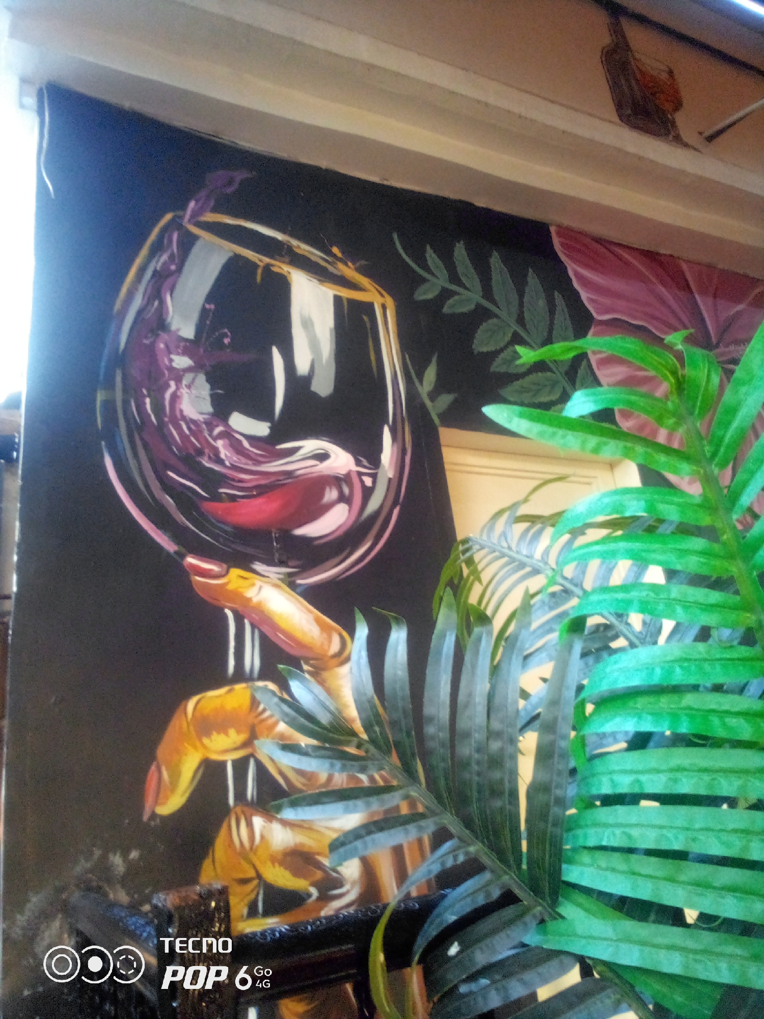 The image depicts a painting on a wall, likely in a commercial establishment, featuring a stylized rendering of a hand holding a wine glass filled with red liquid. The painting's color palette is dark, with contrasting vibrant colors for the hand and the wine. In the foreground, artificial green foliage obscures a portion of the artwork. The bottom left corner of the image contains a watermark with "TECNO POP 6 GO 4G". The broader environment suggests an indoor setting, possibly a restaurant or bar, given the artwork. Without additional visual cues or context, the specific location within Howayi, Nigeria, cannot be determined.