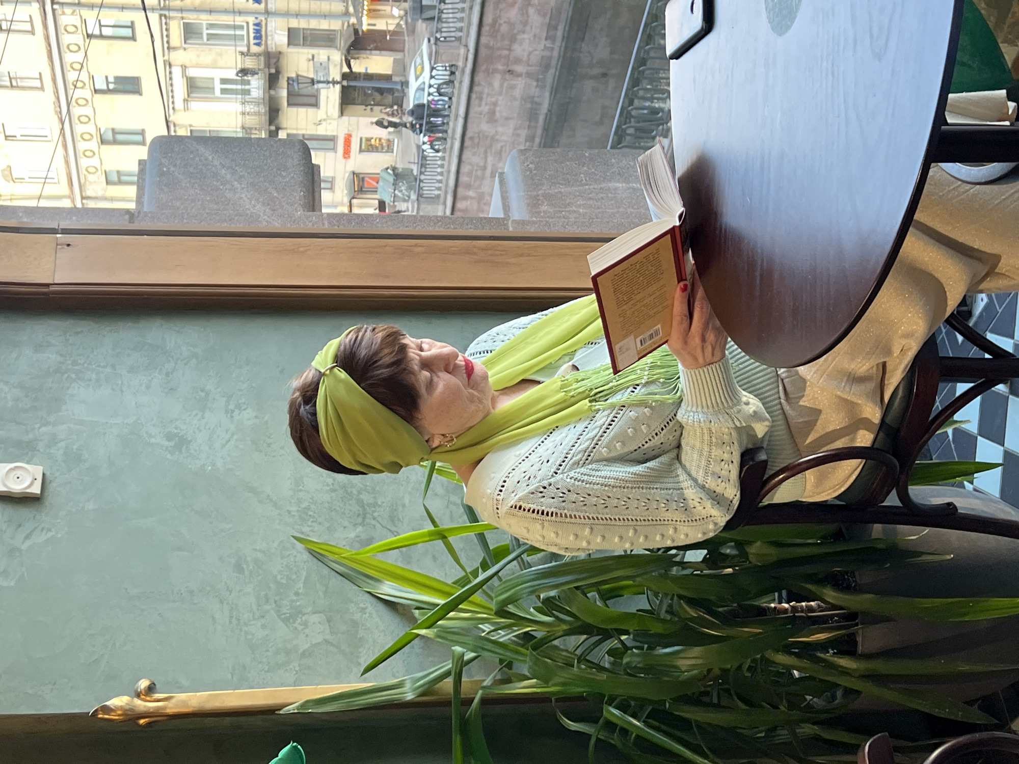 A woman with red hair is seated at a circular wooden table, reading a book with a red cover. She is wearing a light green, lacy sweater and a matching headscarf and scarf with tassels. The table is partially obscured by the book she is reading. A potted plant with long, green leaves sits at the base of the table.
Through the window behind her, buildings in Saint Petersburg, Russia can be seen. There is a canal with a bridge and figures of people walking across. The architecture is predominantly pale yellow. The lighting appears to be natural daylight.