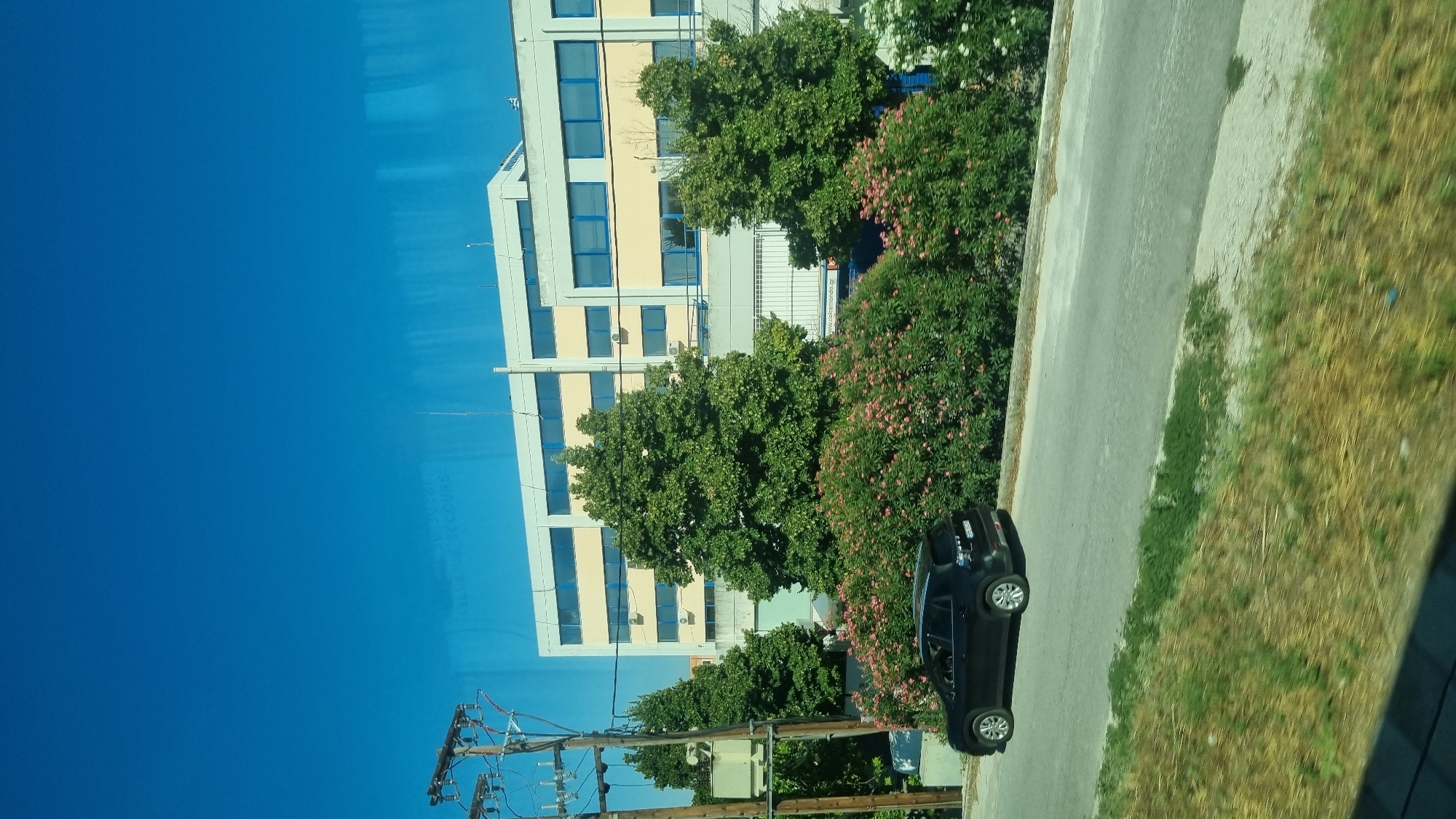 A black SUV is parked on the side of a paved road in Larissa, Greece. The SUV is positioned on the left side of the road, facing right. Behind the SUV, a row of flowering bushes with pink and white blossoms lines the street. Tall, lush green trees are interspersed among the bushes. In the background, a large, modern building with light-colored walls and blue-framed windows stands. A utility pole with wires is visible to the left of the building. The sky is clear and bright blue. The scene appears to be an outdoor view from a moving vehicle.