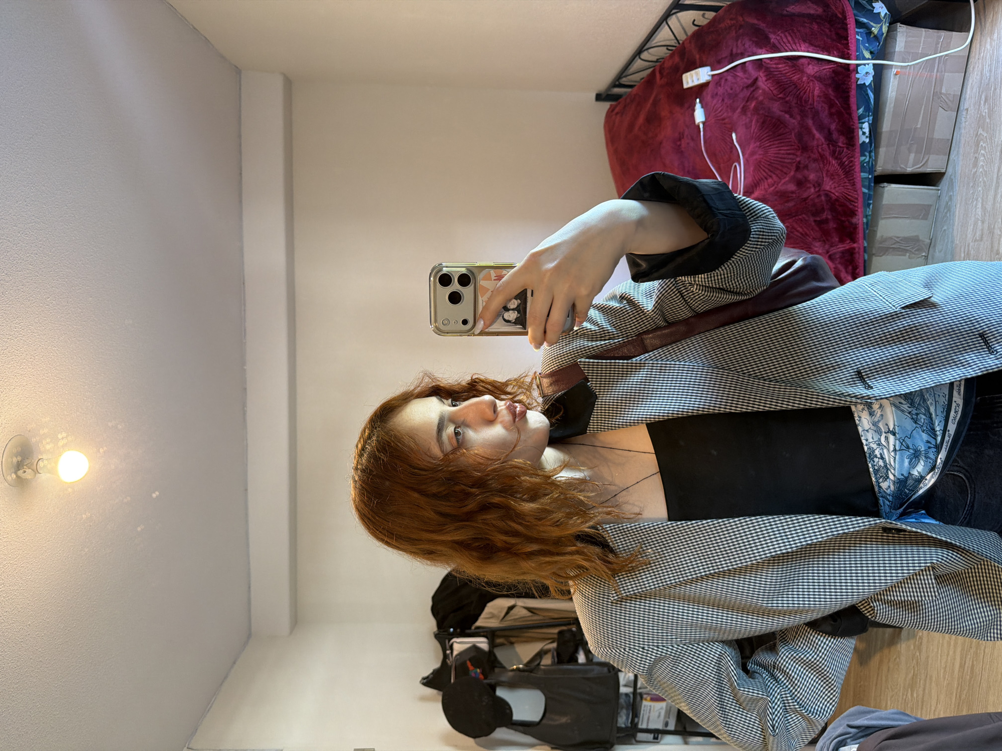 A young woman with long, wavy auburn hair is taking a mirror selfie in what appears to be a bedroom or personal living space in Strasbourg, France. She is looking towards her phone, which she holds up with her right hand to capture her reflection. She has fair skin and her lips are slightly parted in a soft pout.
She is wearing a black camisole or tank top under an open black-and-white checkered blazer or jacket. A dark strap, possibly from a shoulder bag, crosses her chest. Inside her blazer, a glimpse of a light blue patterned fabric, possibly a scarf or jacket lining, is visible. Her lower body and legs are cut off from the frame.
The background shows a simple room setting. To the left, a light-colored wall (likely white or pale gray) features a single bare light bulb that emits a warm glow. To the right, behind the woman's reflection, is a bed with a dark metal bed frame. The bed is covered by a textured dark red blanket, on which a white power strip with charging cables (one white, one black) rests. Behind the bed, against the wall, several brown cardboard boxes are stacked, alongside a piece of dark fabric with a blue floral pattern. The floor, visible towards the bottom right, is light-colored wood. The overall lighting suggests an indoor setting, potentially relying on artificial light. No distinct text is clearly legible on any objects or clothing.