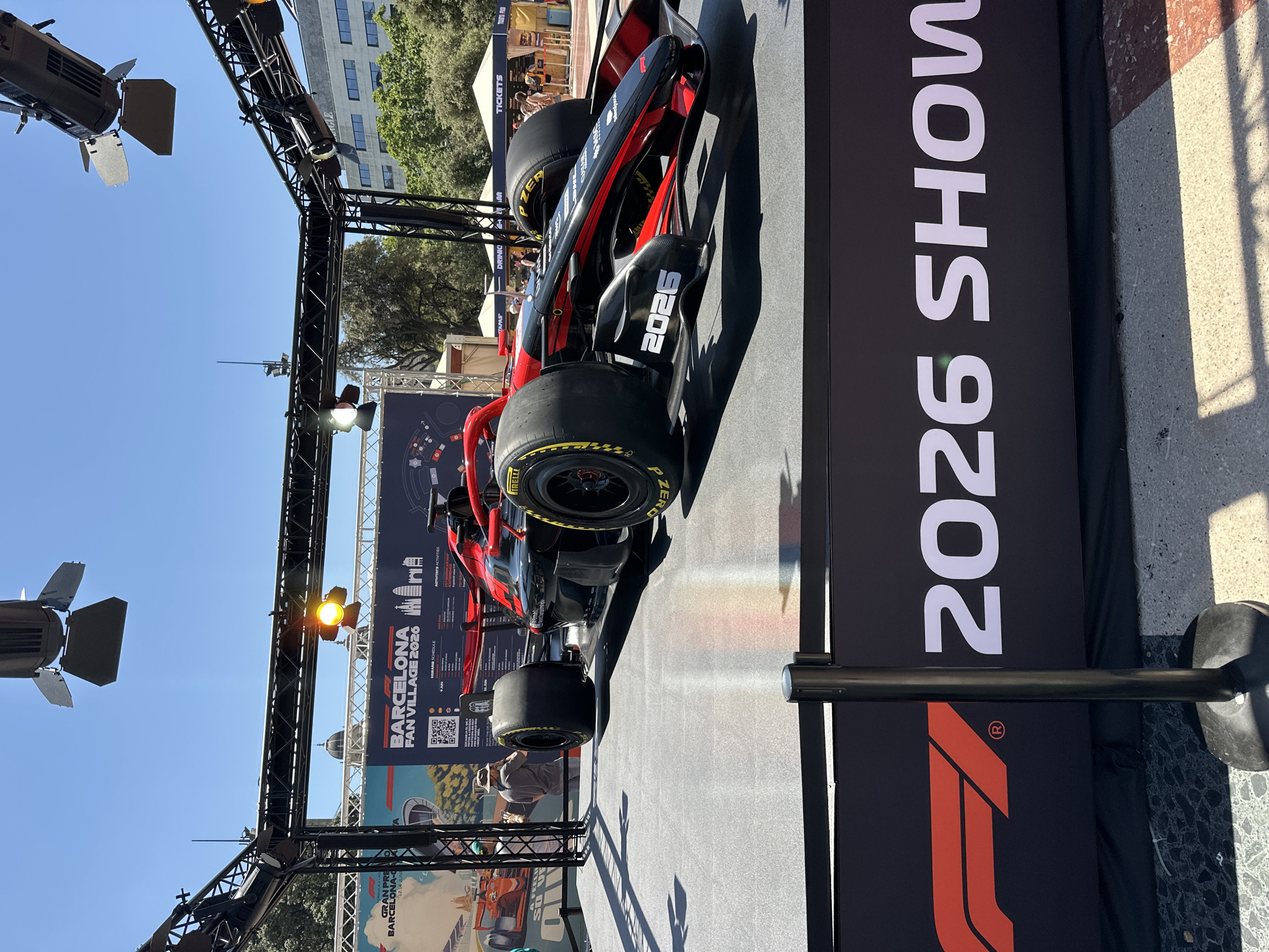 The image shows a Formula 1 car displayed on a raised platform in a public area. The car is black and red, with "2026" visible on its front. In the background, there is a large banner that reads "BARCELONA FAN VILLAGE 2026" and "F1". Several people are visible in the background, some standing and looking at the car, while others are walking or working at booths. The environment appears to be outdoors, with trees and buildings in the distance, suggesting an urban setting. The event is likely a promotional or fan engagement activity for Formula 1 in Barcelona.