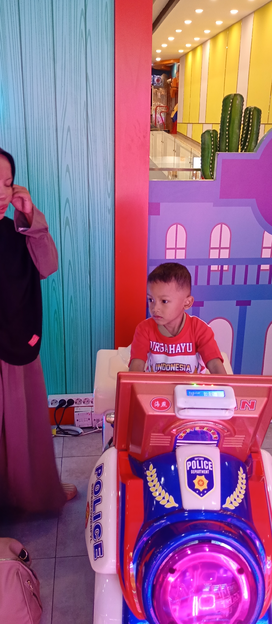 A young boy is sitting in a police-themed ride-on toy. The boy is wearing a red t-shirt with white lettering that reads "URGAHAYU INDONESIA". The toy is blue and red with "POLICE DEPARTMENT" written on it. A woman, partially visible on the left side of the image, is standing near the toy. She is wearing a dark top and a long pink skirt. Electrical outlets and cords are visible on the floor behind the woman. The background features a brightly colored wall with a design that resembles buildings and cacti. The scene appears to be indoors, possibly in an amusement arcade or children's play area. The image quality is moderate, with some blur and noise.
