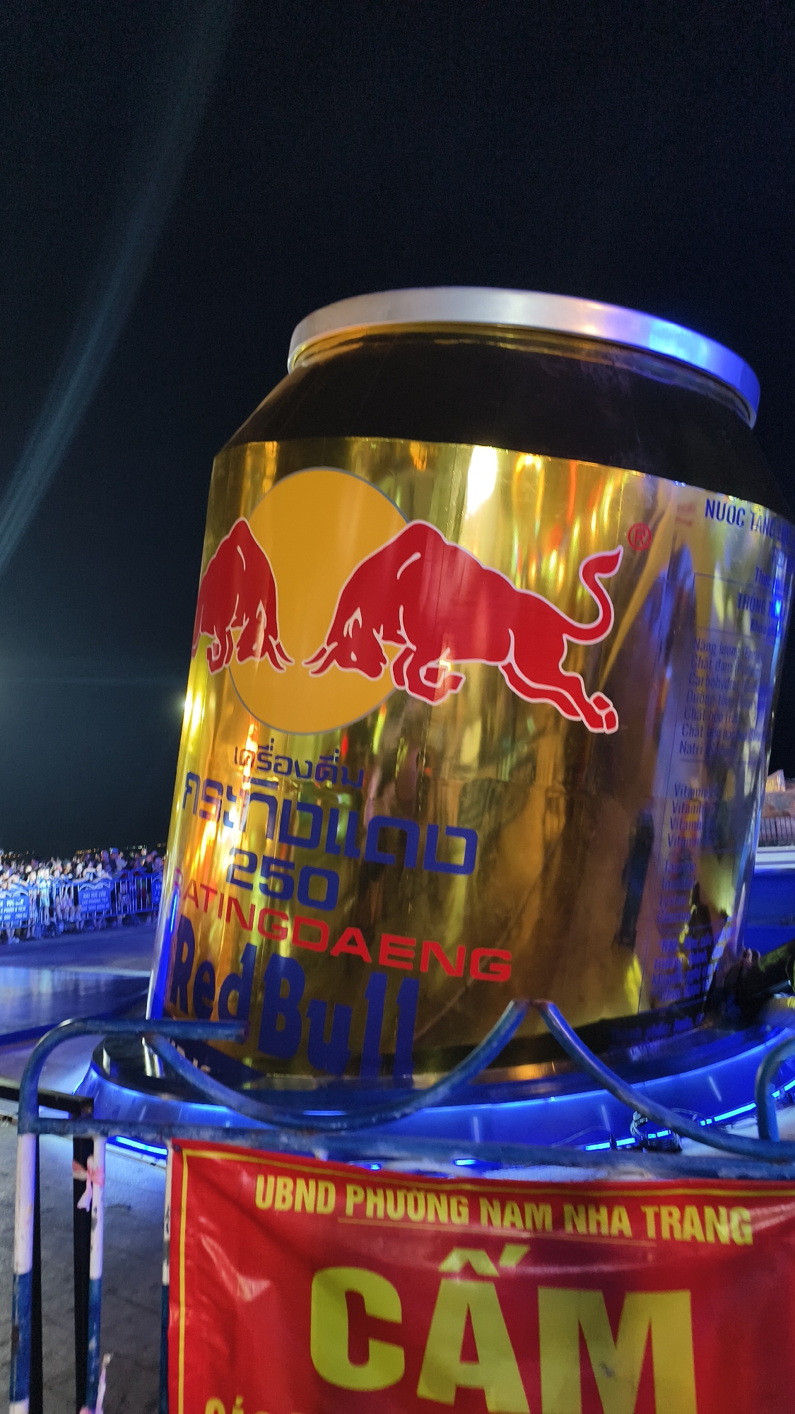 This media file captures a close-up view of a giant, gold-colored Red Bull can, prominently displayed at an outdoor event at night. The can is tilted at an angle, with the iconic red bull logo visible on its surface. The surrounding area is dimly lit, with bright lights, possibly from stage lighting or spotlights, illuminating the can. In the background, a crowd of people can be seen gathered behind blue barriers, suggesting a lively event or festival. A red banner with yellow lettering hangs in the foreground, displaying Vietnamese text: "UBND PHƯƠNG NAM NHA TRANG" and "CẨM" which likely indicates the location and perhaps a welcome message. The overall scene suggests a vibrant nighttime celebration in Nha Trang, Vietnam, with the Red Bull can serving as a central, eye-catching attraction. The text on the can appears to be in multiple languages, including Vietnamese and possibly Thai, with "NUOC TANG" (Vietnamese for "Energy Drink") visible, along with nutritional information like "Năng lưong" (Energy) and "Carbohidrat" (Carbohydrate). The presence of illuminated barriers and the dark sky indicate the event is taking place after sunset.