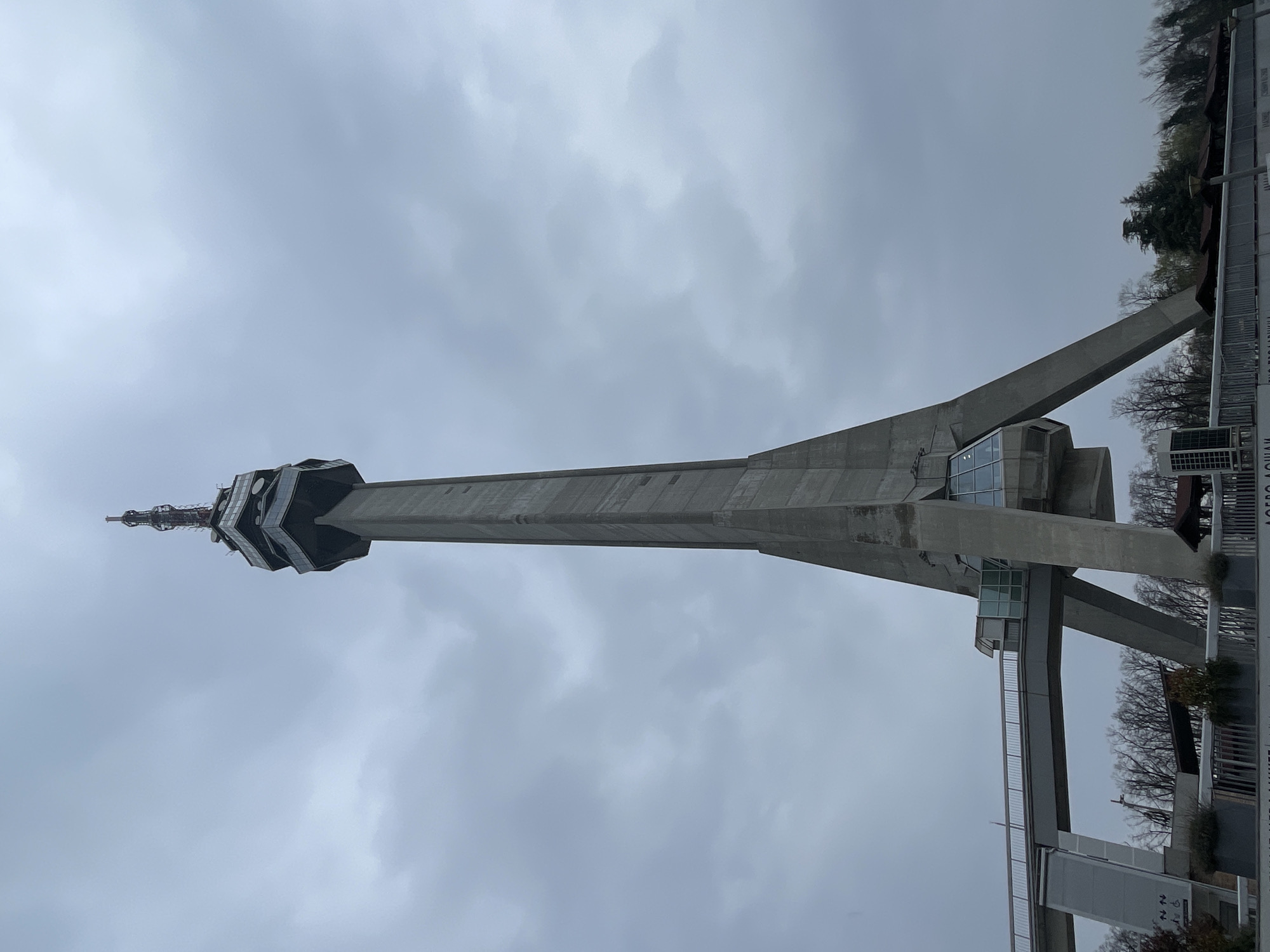 The image shows the Avala Tower in Belgrade, located in the Vozdovac Urban Municipality, Serbia. The tower is constructed of reinforced concrete. The top of the tower has a multi-faceted, glass-enclosed structure. A pedestrian walkway extends from the tower's base. The sky is overcast.