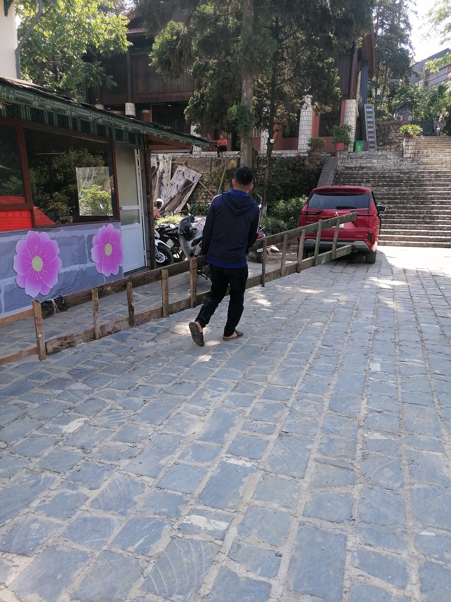 A man in a dark blue jacket and black pants walks on a paved stone surface in Sa Pa Ward, Vietnam. He is facing away from the camera, and a partially constructed wooden fence runs along his left side. Next to the fence is a building with large pink flower decorations and a silver door. To the right of the man is a red car and two motorbikes. Behind the car are stone steps leading uphill. Trees and a larger wooden structure are visible in the background.