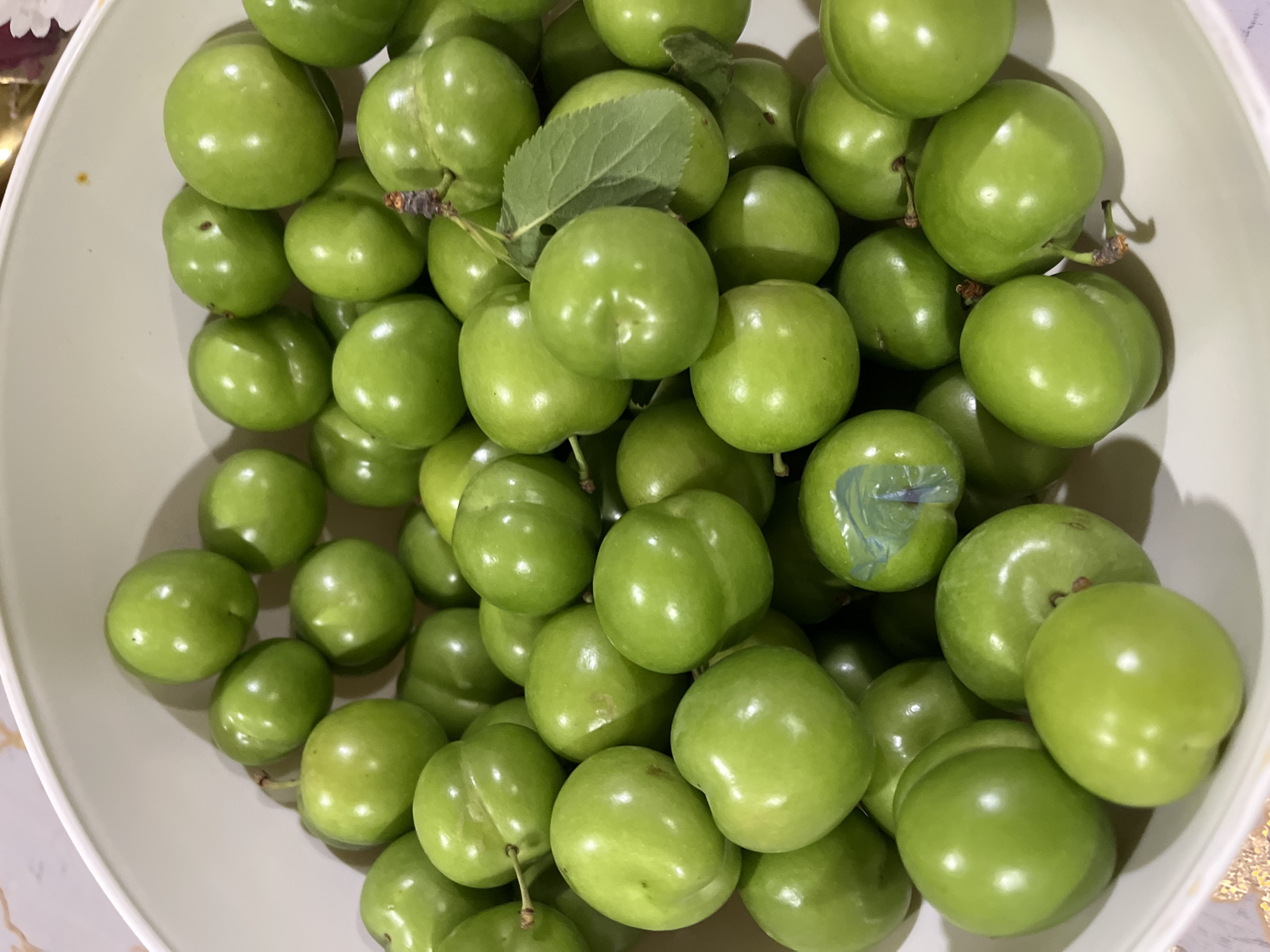A white, circular bowl, partially visible, is filled with a large quantity of small, firm, round to ovate green plums. The plums feature a smooth, glossy, light green skin, with many exhibiting a distinct vertical suture line characteristic of plums. Small brown specks or minor blemishes are visible on the surfaces of some fruits. One plum, situated in the central-right portion of the bowl, retains a short, dark brown stem with a single, small green leaf attached. Another plum, located in the lower-central area, has a small, irregular blue marking on its skin. The fruits are tightly clustered, reflecting ambient light and casting soft shadows within the bowl. The background beyond the bowl is softly blurred. This scene is located in the city of Unknown, Unknown.