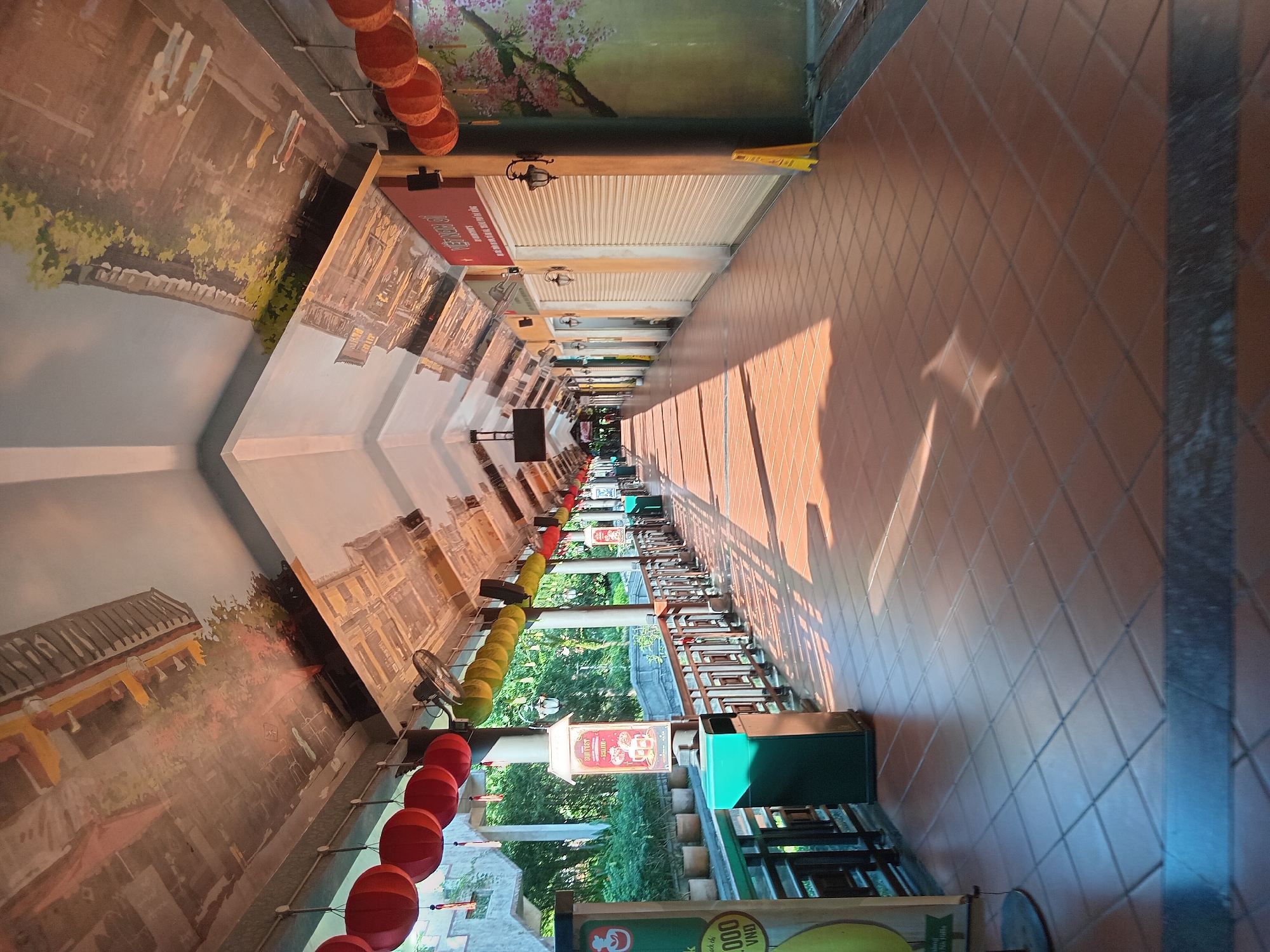 The image shows an outdoor covered walkway in Da Nang, Vietnam, decorated with murals of traditional Vietnamese architecture and a cherry blossom tree. Red lanterns hang from the ceiling on the left side, and yellow lanterns hang on the right. The walkway is paved with brown tiles, and shops with closed roll-up doors line one side. Green trash cans are positioned along the walkway. Sunlight streams through the open sides, casting shadows on the tiled floor. The overall impression is one of a peaceful, possibly commercial, area during daytime.