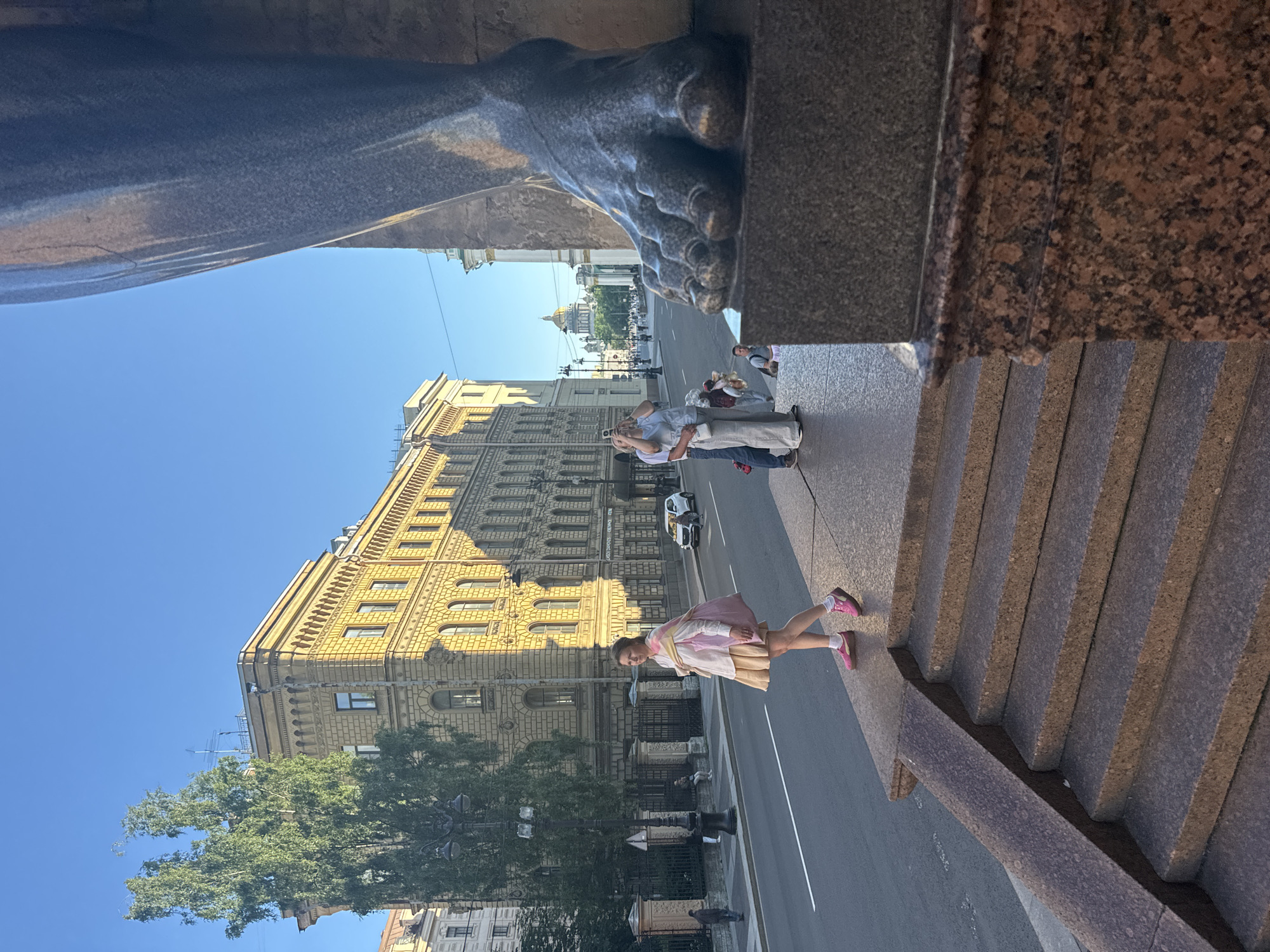 The image captures a daytime scene in Saint Petersburg, Russia, likely on Nevsky Prospekt, near the Kazan Cathedral, as suggested by the architectural style of the buildings and the prominent dome in the background. In the foreground, a large, weathered stone foot of a statue occupies the right side of the frame, casting a shadow. To the left, a woman in a light-colored blouse, a beige skirt, and pink sneakers walks along a granite staircase. She carries a large pink bag over her shoulder. Behind her, a street with a few cars and pedestrians stretches into the distance, bordered by a building with a yellow facade. A couple is visible in the mid-ground, with one person holding a phone, possibly taking a photograph. The sky is clear and blue.