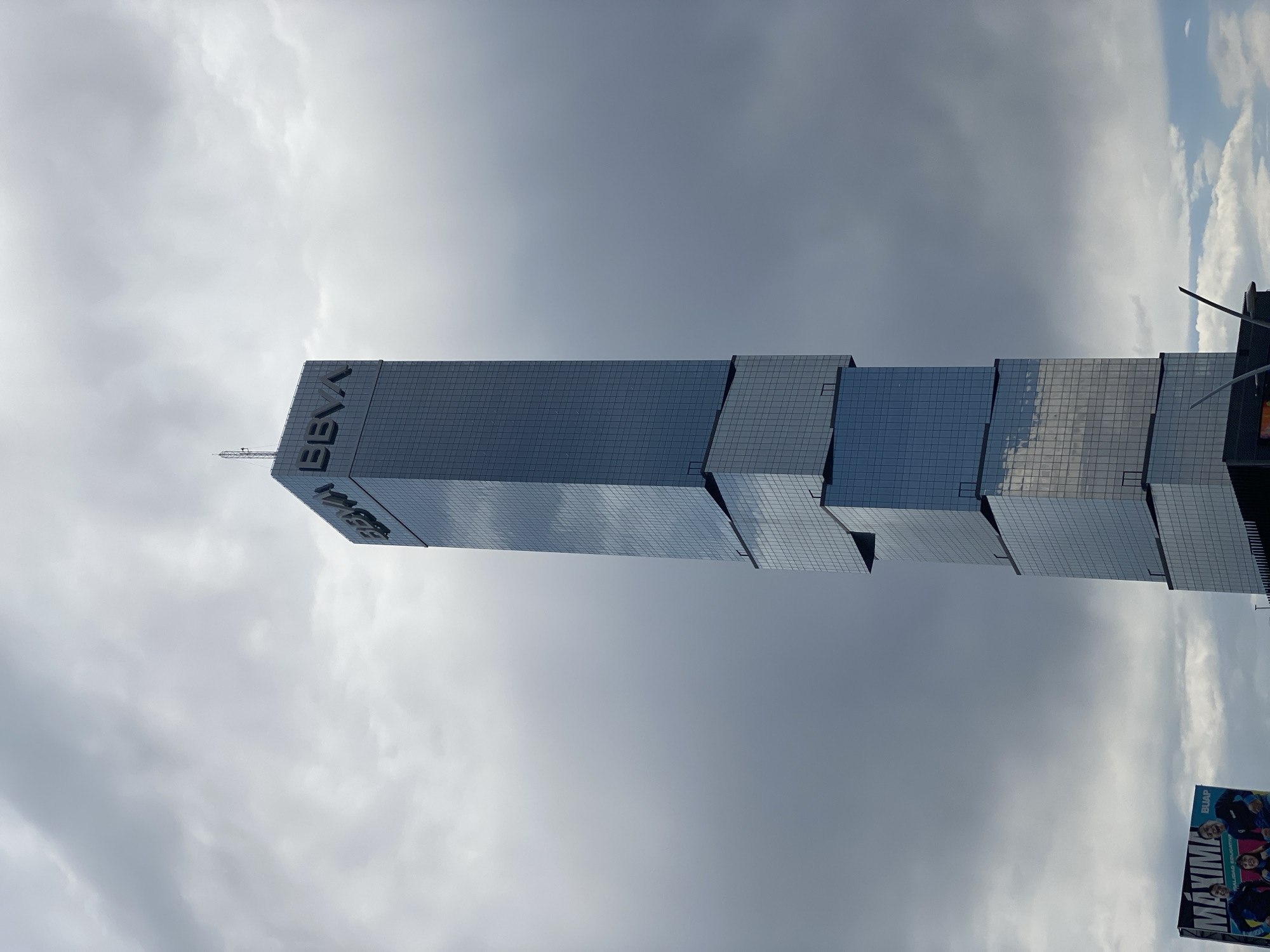The image shows the BBVA Tower in Puebla, Mexico. The tower is a modern skyscraper with a distinctive stepped design and a reflective glass facade. The sky is overcast with grey clouds, and a portion of a billboard is visible in the lower right corner.