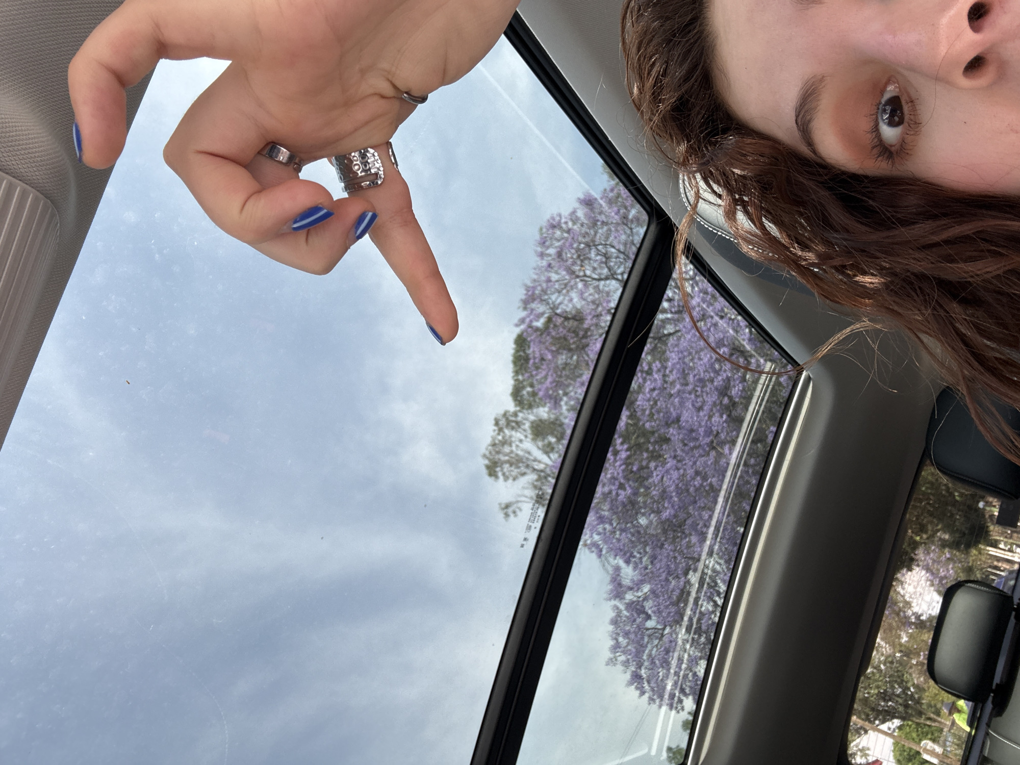 The image shows a partial view of a person inside a car, likely in Mexico City based on the presence of Jacaranda trees in bloom, which are common in the city. The person is looking up towards a sunroof with their right eye visible. Their brown hair is visible along the right side of the frame. They have blue fingernail polish on their left hand and are wearing multiple rings. The left hand is positioned above the sunroof, with the index finger pointing downwards toward the glass. Through the sunroof, the sky is visible with light clouds. The tops of Jacaranda trees with purple blossoms are also visible. The interior of the car includes the headliner and trim along the sunroof frame. Outside the car, reflected in the glass, are other trees and street-level scenery.