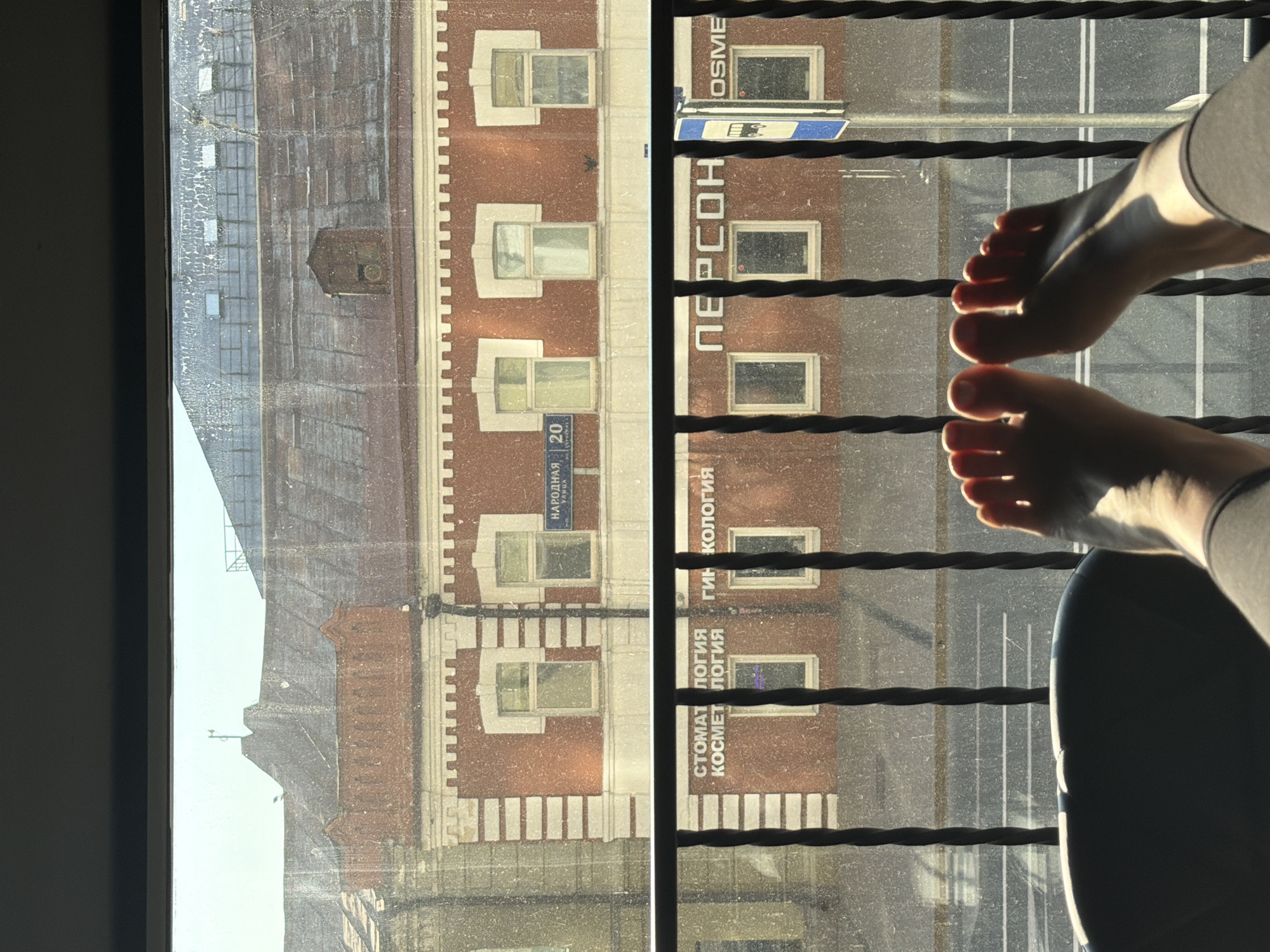 The image shows a view from a window or balcony overlooking a city street. In the foreground, two bare feet are visible, resting on a dark surface, likely a chair or sofa. The feet are illuminated by sunlight coming from the left, casting shadows. A black metal railing is in front of the feet, with twisted vertical bars.
Beyond the railing, a building with red brick facade and white decorative elements is visible. The building has multiple windows, some of which are reflecting the sky. A street sign with Russian text, "НАРОДНАЯ 20," is mounted on the building. Below this, on the lower part of the building, there are signs with text that appears to be in Russian: "СТОМАТОЛОГИЯ," "КОСМЕТОЛОГИЯ," and "ПЕРСОН." A bus stop sign with a bus icon is also visible to the right.
The street below the building is paved and has yellow and white lines, indicating traffic lanes. The sky above is bright, suggesting daylight. The glass of the window or balcony appears dirty, with streaks and smudges.
There is no visual information in the image to suggest the location is in Укромновское сельское поселение, Ukraine. The presence of Russian language signage indicates a location where Russian is spoken.
