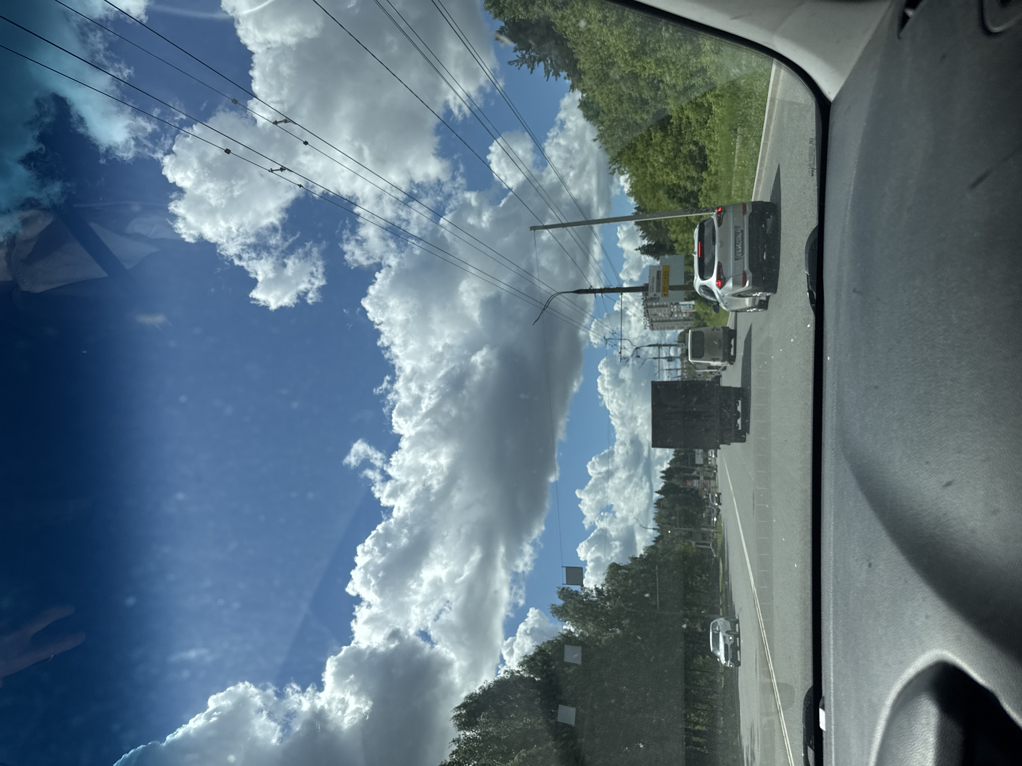 The image, taken from inside a vehicle, shows a view out the driver's side window on a road. The sky is predominantly blue with white cumulus clouds. Overhead power lines are visible against the sky. The road has multiple lanes of traffic with several cars in front of the vehicle. Trees and greenery line the side of the road. Signage is visible, but the text is not legible. The location is possibly Izhevsk, Russia, but without specific identifying landmarks, this is not confirmed. No people are visible.