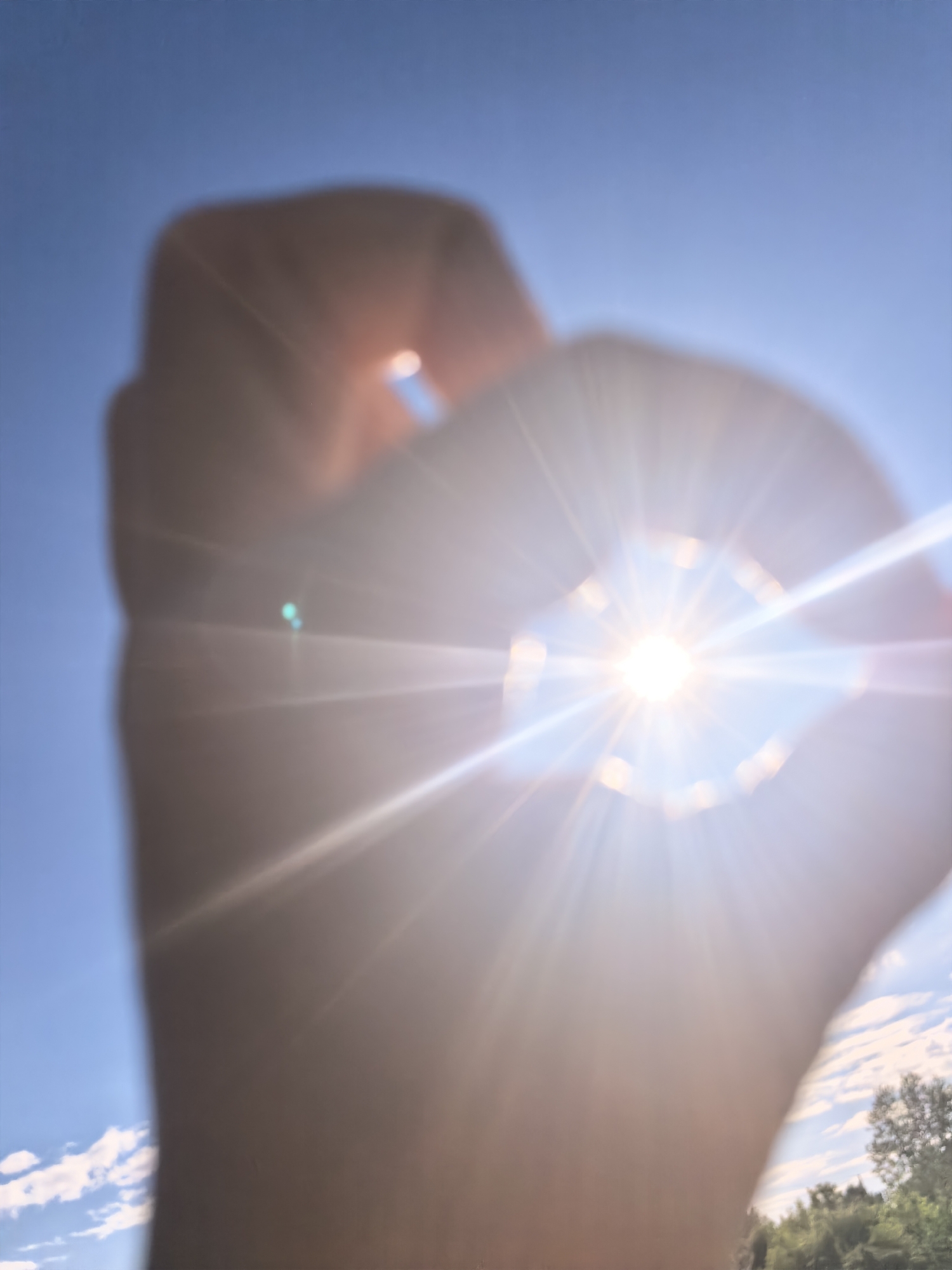 A person's hand, out of focus, is held up in front of a bright sun, creating a starburst effect. The sun is in the center of the frame, and its light flares out in all directions, overwhelming the image. The sky is a clear blue, with a few wispy clouds visible in the distance. The background shows a blur of green trees and more clouds. The image appears to be taken outdoors during the day.