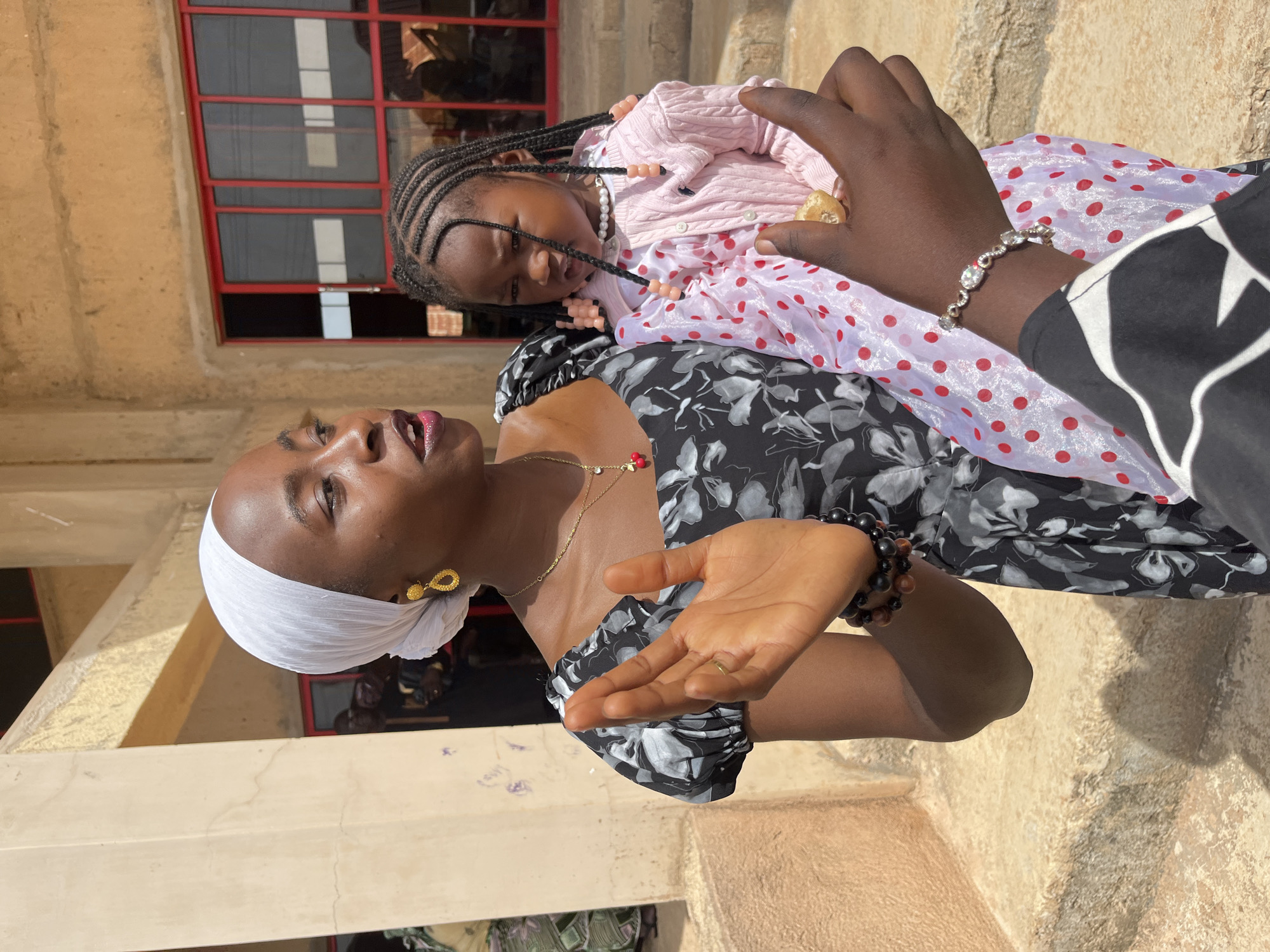 A medium shot captures a woman in a white headscarf and a black and gray floral dress holding a young child. The woman's left hand is raised with fingers splayed, and her right arm cradles the child. The child, dressed in a pink cardigan over a white and red polka-dot dress, looks slightly to the right with braided hair adorned with pink beads. The backdrop is an outdoor setting with a building featuring red-framed windows and textured walls. The ground appears to be made of concrete or stone steps. The scene is illuminated by natural light, casting shadows.