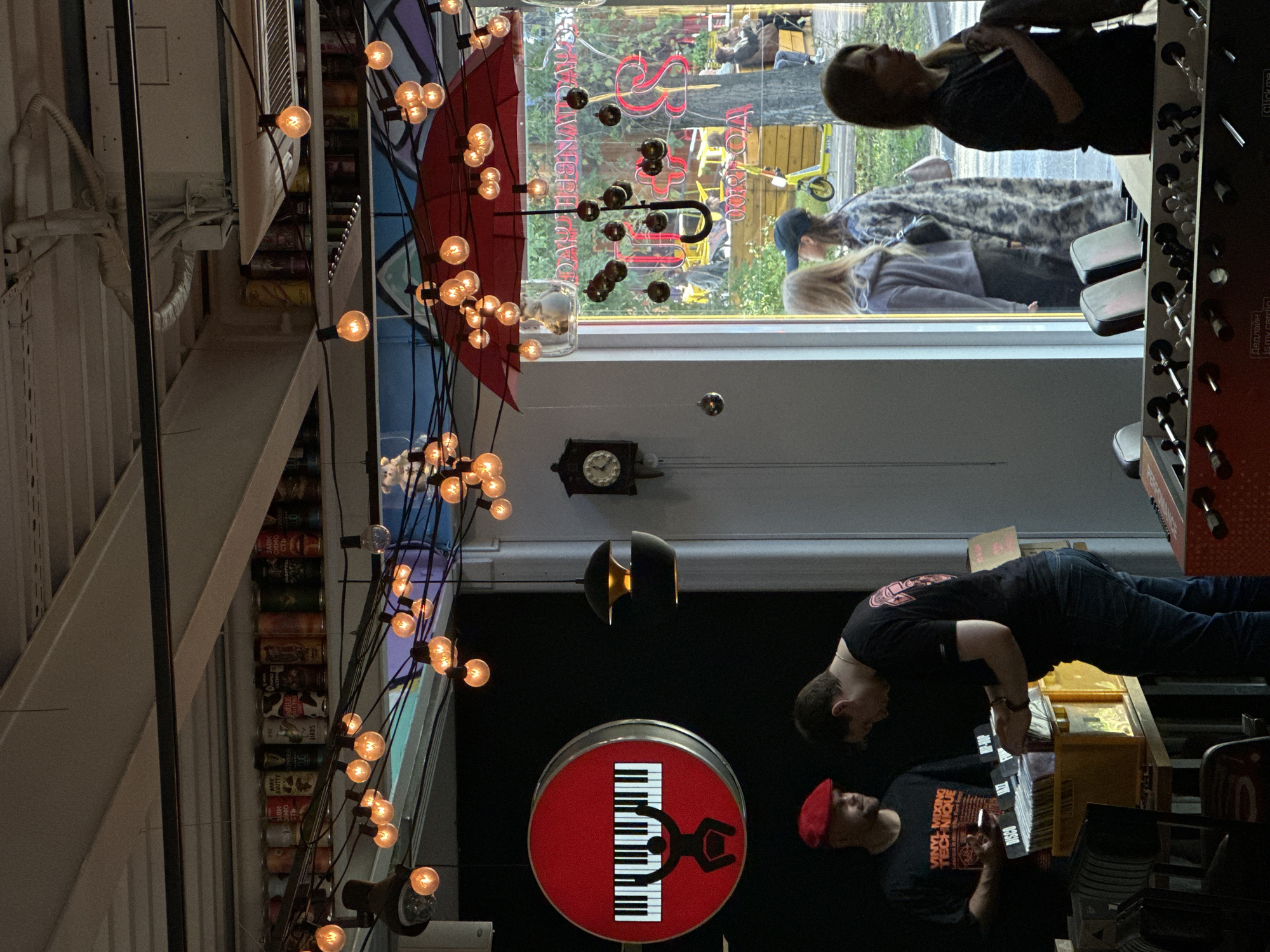 The image depicts the interior of a record store in Khimki, Russia. Two men are visible in the foreground, one wearing a red beret and a t-shirt with "VINYL MIXING TECHNIQUE" printed on it, and the other in a black t-shirt. They are standing behind a table with crates of vinyl records. The store is decorated with numerous string lights and a red umbrella with lights hanging from it. A clock hangs on the wall. Through a large window, several people are seen outside, and a yellow scooter is parked. The scene suggests a casual, community-focused atmosphere, possibly a gathering or event related to music.