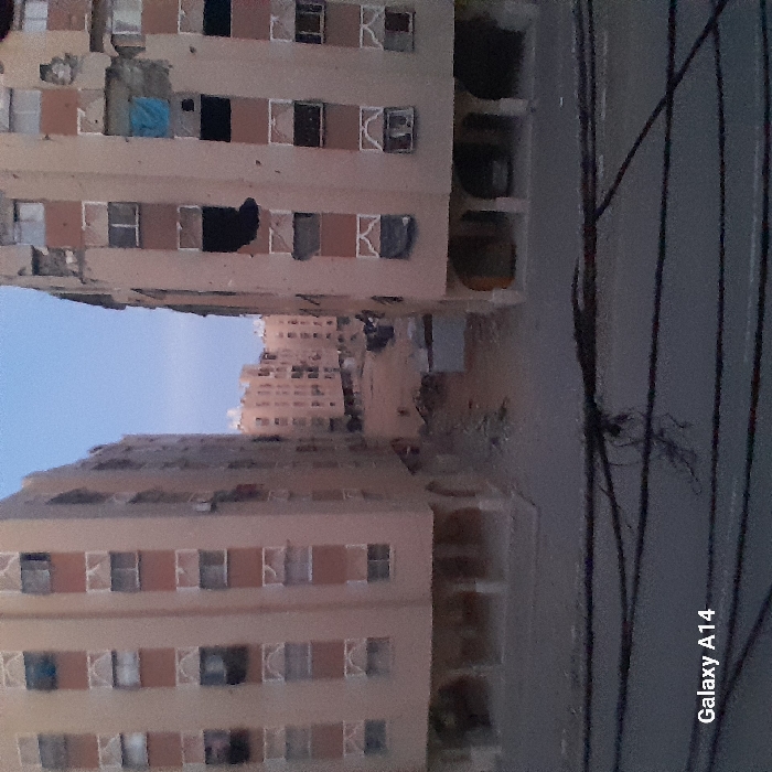 The image presents an urban residential scene, likely captured from a building or elevated vantage point looking across a street or alleyway towards other structures. The overall impression is one of dense habitation in a somewhat aged urban environment.
In the foreground, dominating the left side, is a multi-story apartment building with a light beige or pale brown facade. Numerous windows are visible, many with white frames. Some windows appear to be open, and various items, including blue and dark-colored fabrics, are seen hanging from windows or small balconies, suggesting laundry drying. Parts of this building show signs of wear, with some discolored or damaged sections. Distinctive white, stylized "K" shapes, possibly decorative window guards or parts of railings, are visible near many windows.
Across a visible gap, which appears to be a street or unpaved area, another similar apartment building is situated on the right side of the midground. This building shares the same light color scheme and multi-story design with many windows. Below this building, at what would be ground level, are open-sided structures that resemble garages, storage areas, or open balconies, with some vague objects, possibly pots or containers, visible within.
Further in the background, behind these primary structures, more buildings are visible, forming a dense urban backdrop. They are less distinct due to distance and slightly hazy conditions. The sky above these distant buildings and to the upper left is a clear, pale blue, indicating either early morning or late afternoon/dusk, given the soft, ambient lighting on the buildings.
On the right side of the image, stretching vertically, are several dark, thin lines that appear to be utility wires or cables. Some tangled dark material, possibly more wires or dead vegetation, is seen interwoven among these lines.
No people are distinctly visible, and no specific activity is clearly captured beyond the general presence of residential life implied by the buildings and hanging laundry. The overall atmosphere is quiet and still.
At the bottom right corner of the image, the text "Galaxy A14" is visible, indicating the model of the smartphone used to take the picture.