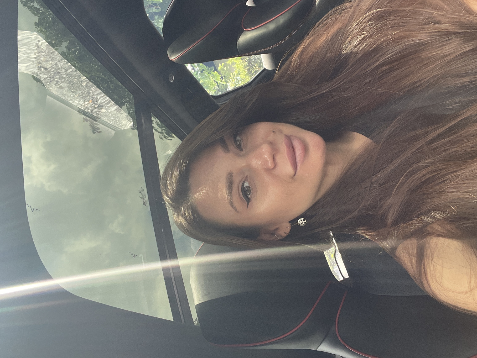 A woman with long brown hair is sitting in a car, presumably driving, and facing the camera. She has a fair complexion, and her hair is brushed to one side. She is wearing earrings. The car has a black interior with red trim on the seats. The sky is visible through the car's sunroof. The location is Unknown, Unknown.