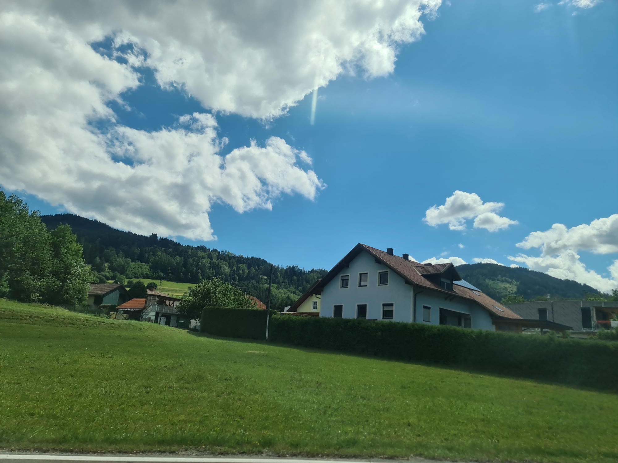This is a daytime, eye-level, exterior shot of a rural scene in Sankt Lorenzen bei Scheifling, Austria. The foreground is dominated by a lush, green, mowed lawn that slopes gently upwards. In the middle ground, a light blue, two-story house with a dark red, sloped roof is visible. It has several windows on its facade. Adjacent to this house, further back and to the left, are other buildings with red roofs. A neatly trimmed green hedge runs along the property line in front of the houses. The background features a densely forested, verdant mountain slope under a bright blue sky filled with scattered, fluffy white clouds. A faint sunbeam is visible cutting through the clouds.