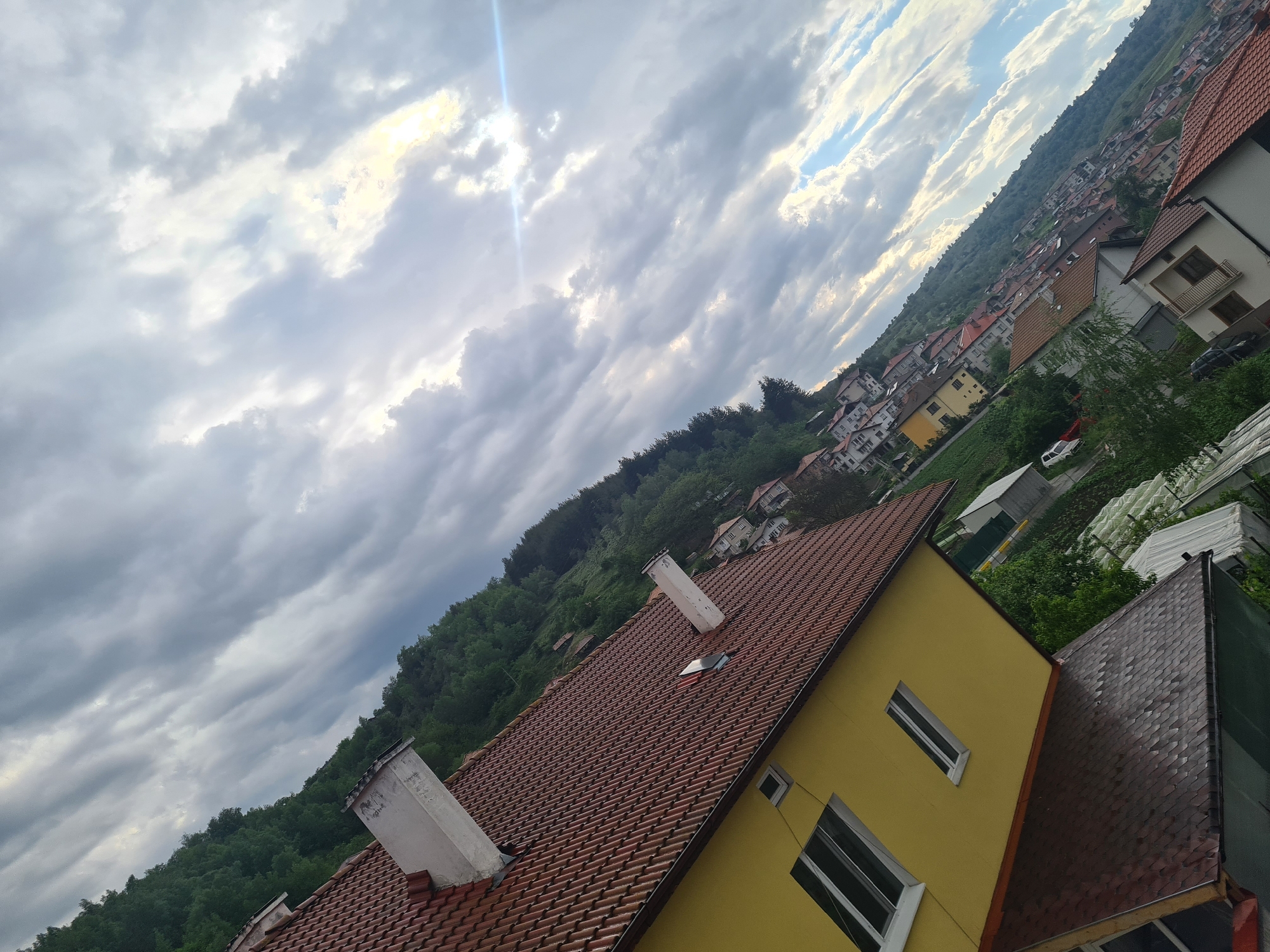 The image, taken at an oblique angle, presents a view in Kochan, Bulgaria, focusing on residential architecture against a backdrop of sky and forested hills. A yellow house with a red tile roof and two white chimneys dominates the foreground. The house has at least two windows visible on its facade. Adjacent to the yellow house is a building with a dark-colored shingle roof.
Beyond the houses, a densely populated area of similar residential buildings spreads up a hillside, interspersed with trees. A forest covers a larger hill in the middle distance. Above, a sky filled with layered grey and white clouds is visible, with hints of sunlight breaking through in some areas. There are no people visible in the image.