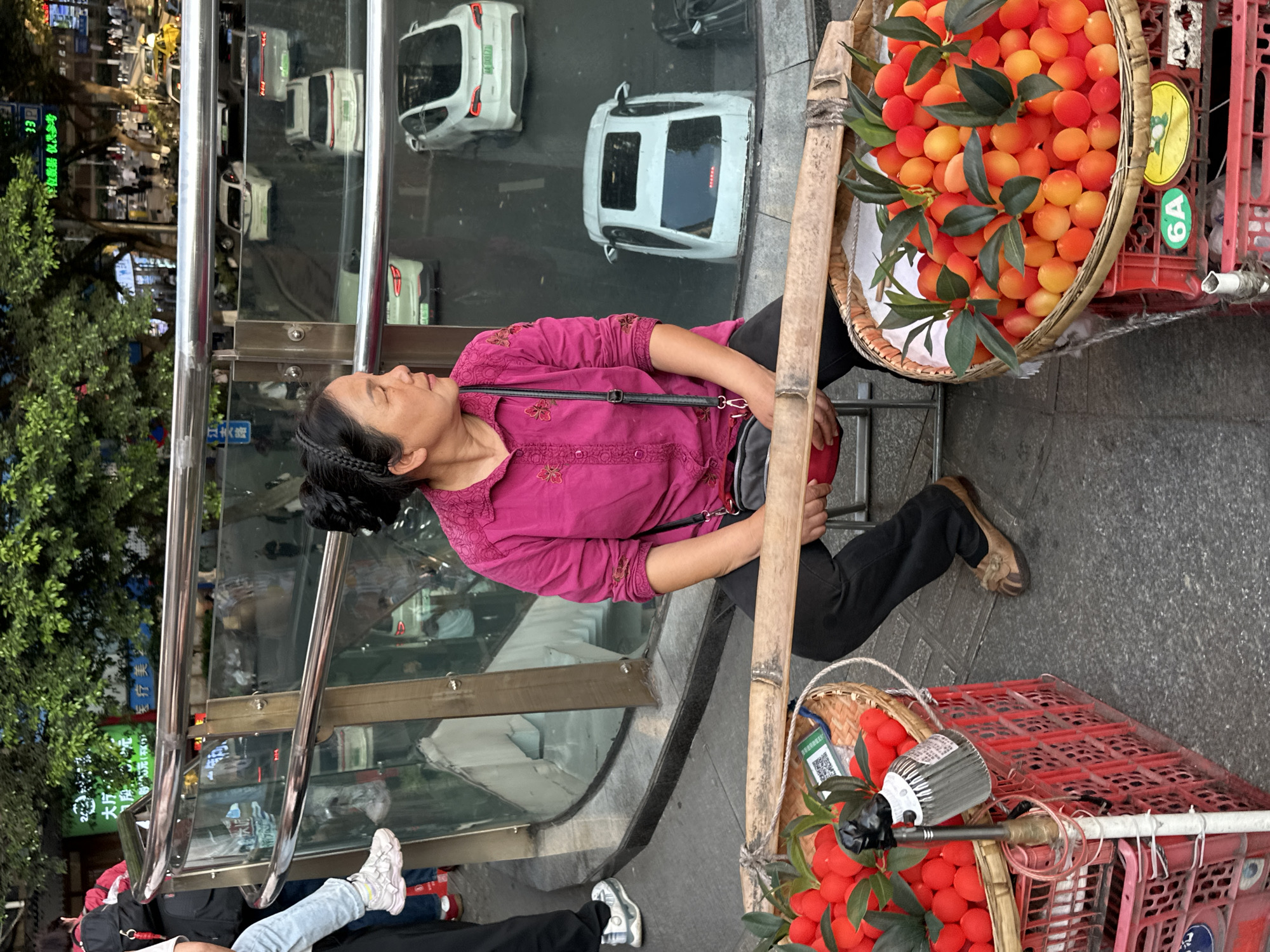 Here is a detailed description of the image:
The image appears to be taken at a slightly tilted angle outdoors in the Yuzhong District of China. A vendor sits next to a bus stop or a similar enclosed waiting area. The vendor is an Asian woman with black hair pulled back into a bun and adorned with a dark headband. She is wearing a magenta long-sleeved shirt and black pants, along with tan shoes. She has a black crossbody bag. She seems to be resting or sleeping, leaning back against the glass wall of the bus stop.
The vendor is selling fruit, which is displayed in two baskets. One basket, placed on top of a red plastic crate, is filled with round, orange fruits, possibly Kumquats, complete with leaves. The second basket, also placed on a red plastic crate, contains similar round, red fruits. A bamboo pole is leaning against the baskets, presumably used for carrying them.
In the background, through the glass wall of the bus stop, there is a reflection of the city street, with cars and trees visible. There are pedestrians walking around the area, some of whom are partially visible in the foreground. Street signs with Chinese characters are visible in the background. One street sign says "江大路" which is "Jiangda Road." There's also text on the sign saying "大厅 元(不)医疗美" which translates to "Hall Yuan (not) medical beauty." The street is likely a bustling area with shops and vehicles.
The general environment is an urban setting, with a street and pedestrian traffic.